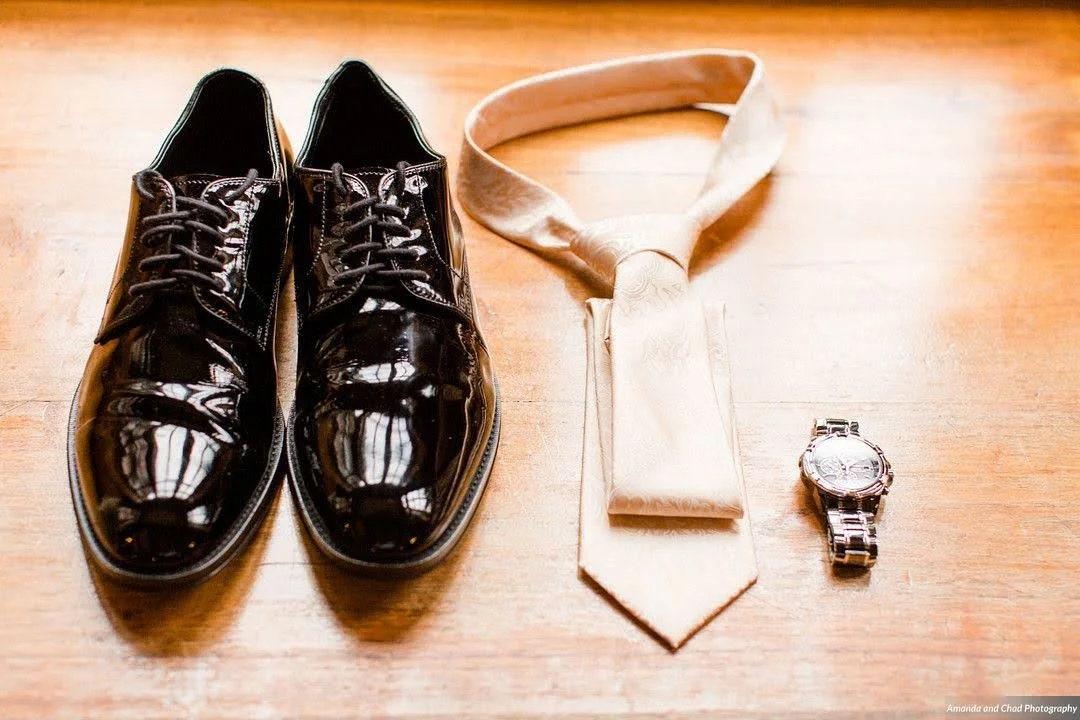 Groom detail flat lay with black dress shoes, a tie, and a wristwatch on a wooden surface.