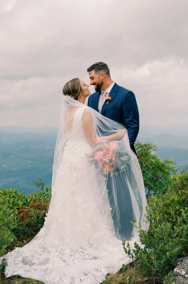 Bride and groom kiss outdoors with mountain views, the bride’s veil flowing in the breeze.