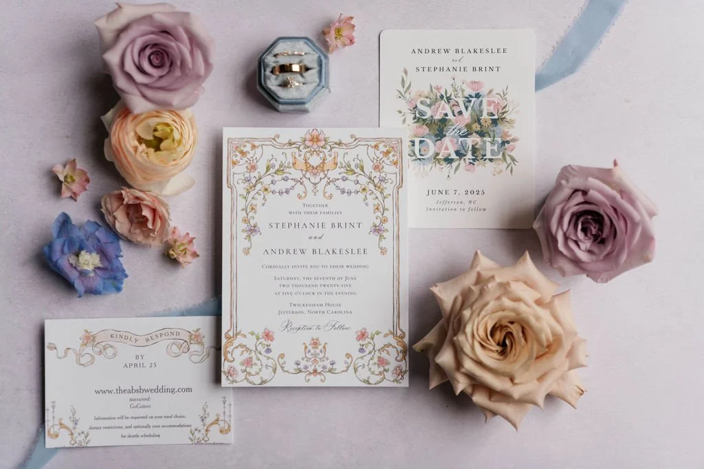Wedding stationery flatlay with floral invitations, rings, and roses on a pastel backdrop.