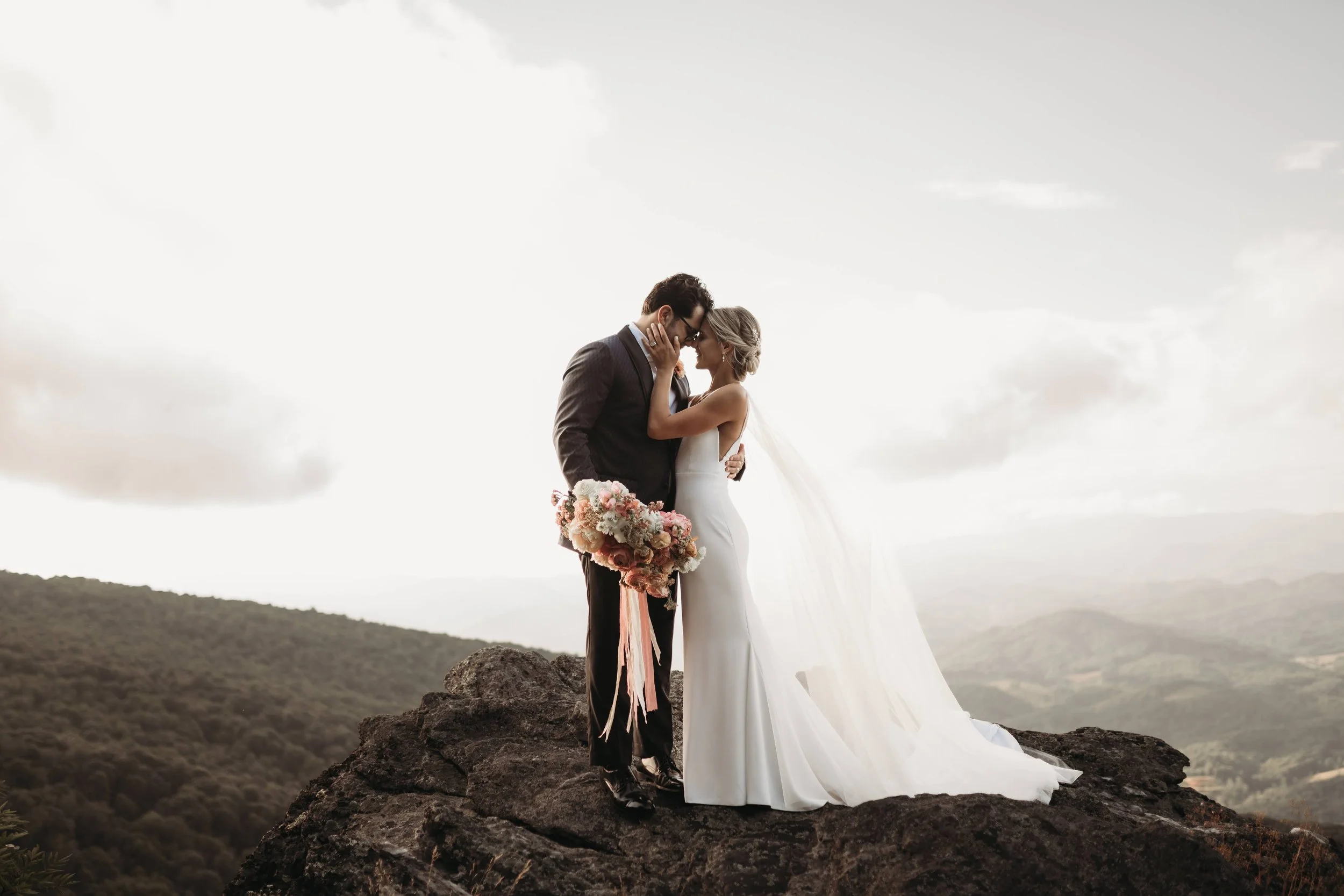 Bride and groom embracing on a rocky mountain cliff with dramatic clouds, valley views, and a flowing veil.