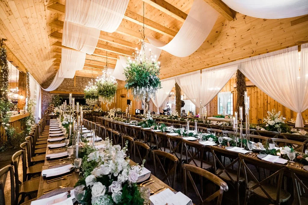 Reception hall featuring long wooden tables and stunning chandeliers adorned with lush green foliage.