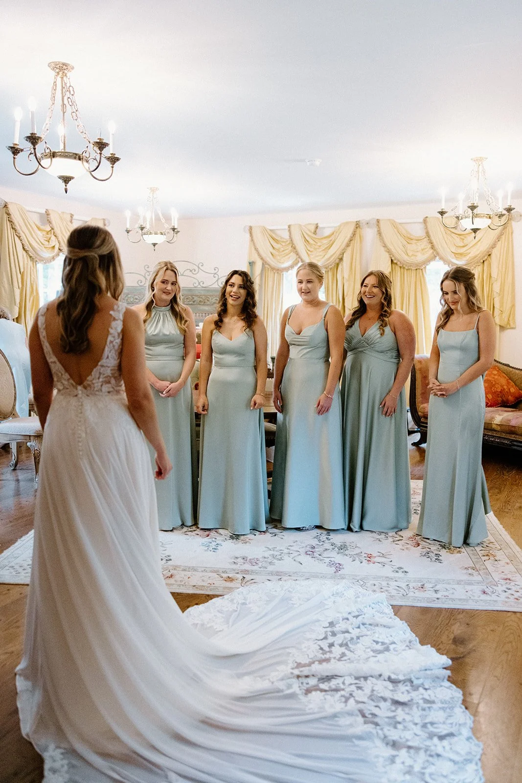 Bride revealing her wedding dress to bridesmaids in light blue dresses inside an elegant getting-ready room.