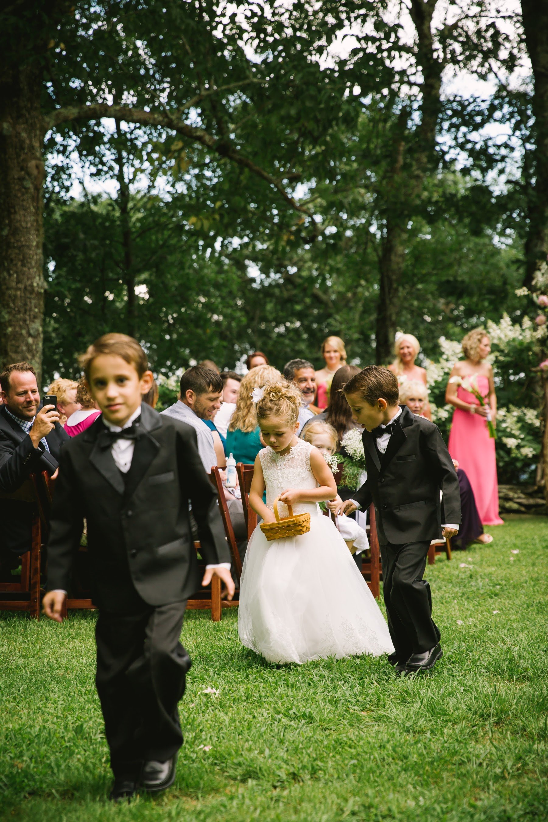 Children walk down the aisle outdoors, with a flower girl holding a basket and boys in tuxedos beside her.