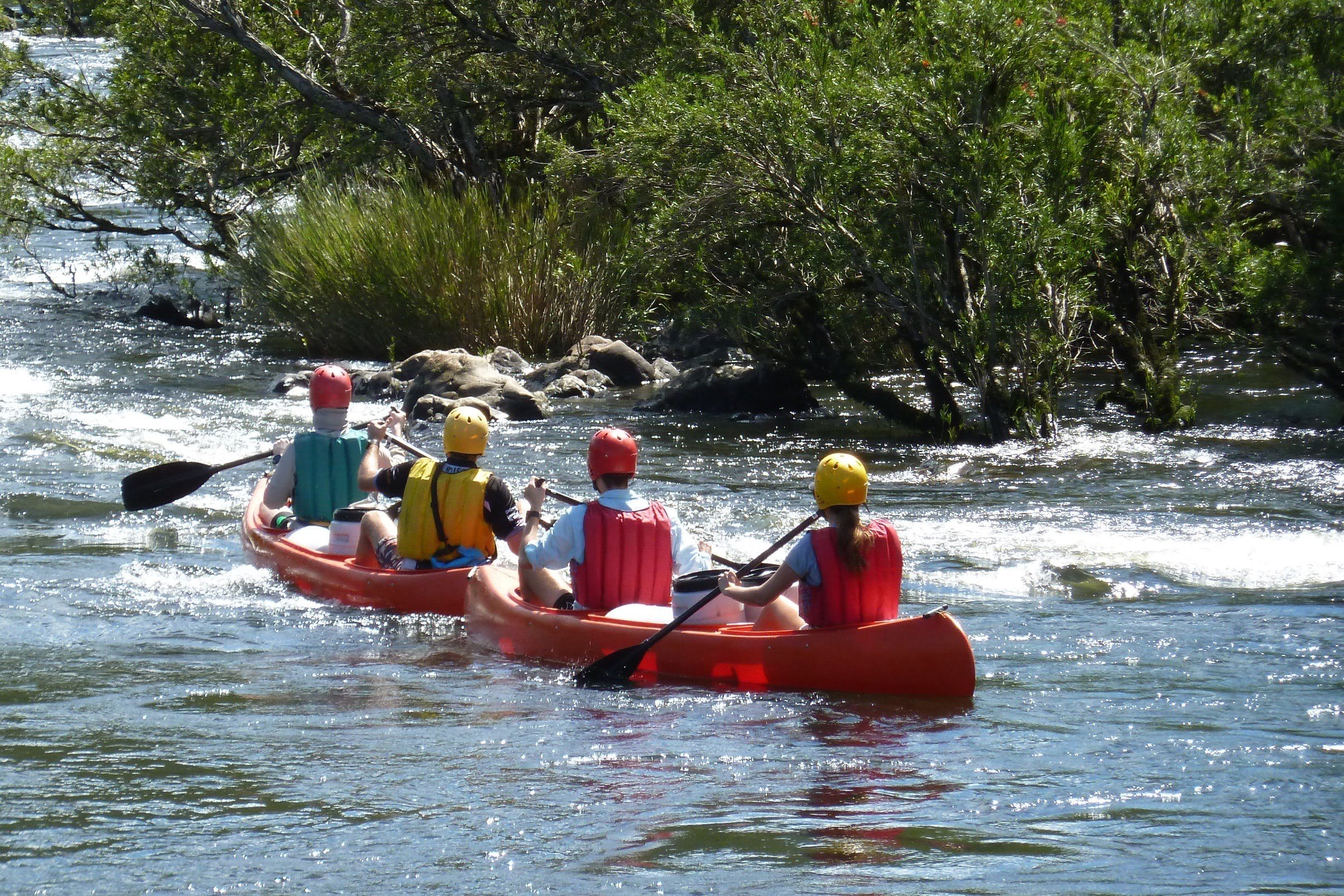 Group paddling in inflatable boats through shallow river rapids wearing helmets and life jackets.