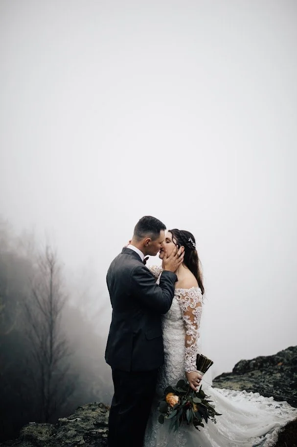 Bride and groom kissing on a rocky overlook in heavy fog, bride holding a bouquet.