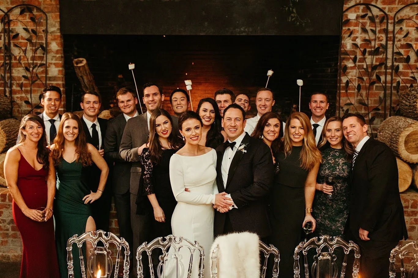 Group portrait of wedding guests in formal attire posing in front of a large fireplace indoors.