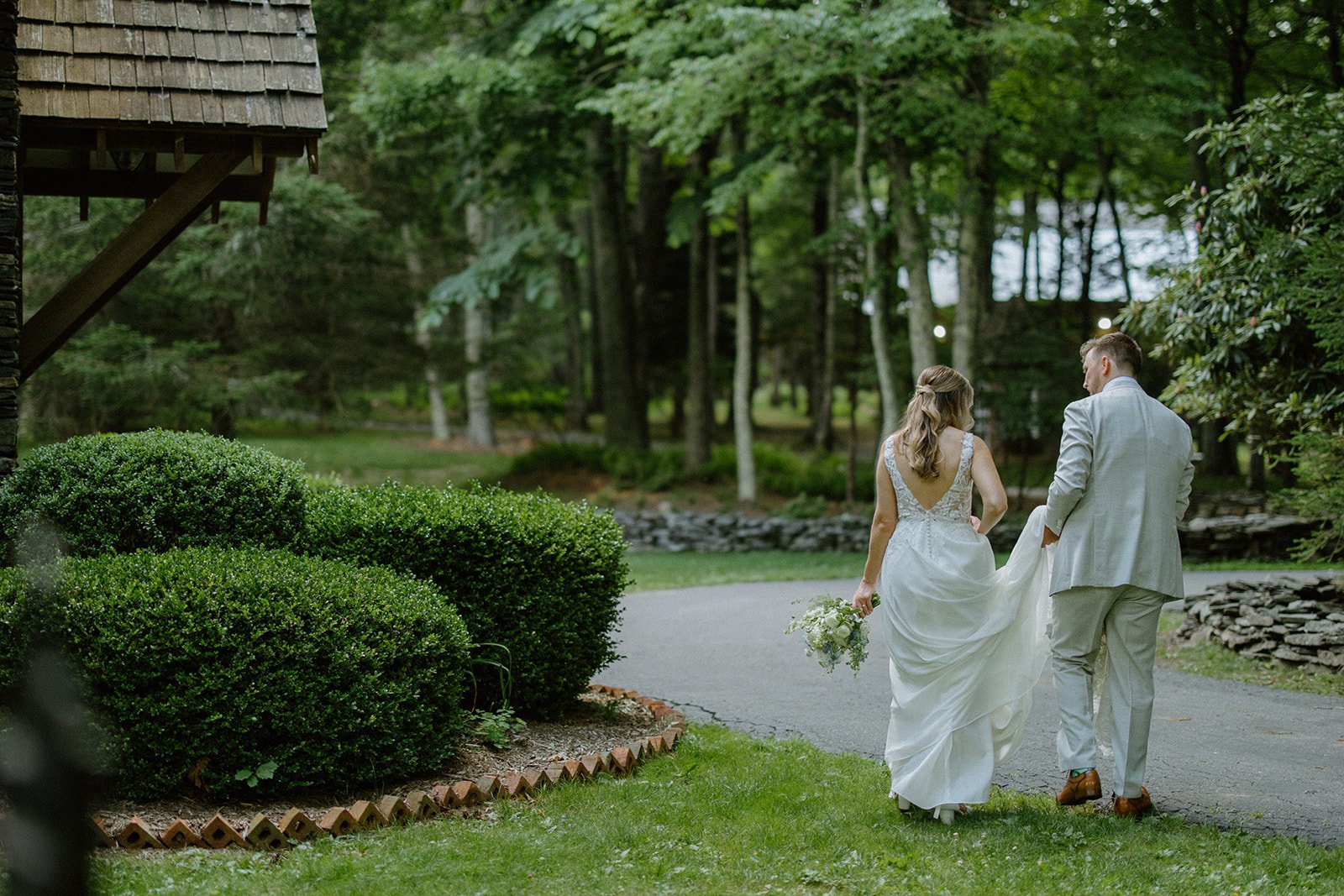 Newlyweds walking hand in hand through a wooded estate garden after their wedding ceremony.