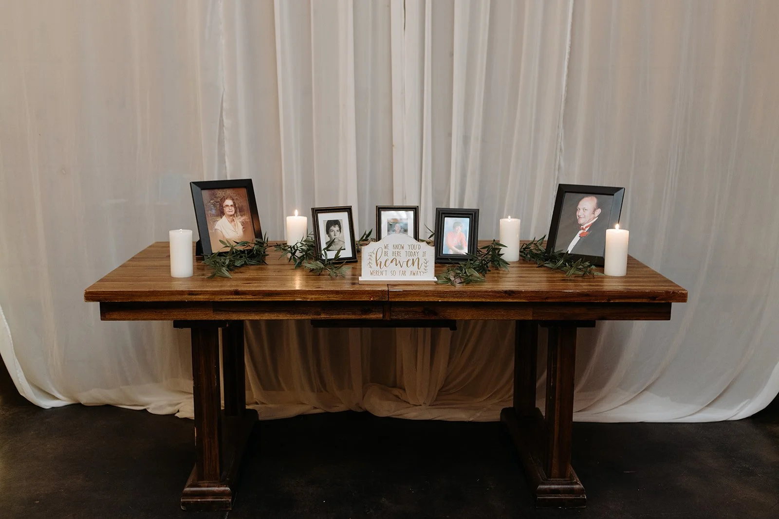 Wedding memorial table with framed photos, white candles, greenery, and a sign displayed in front of a white draped backdrop.