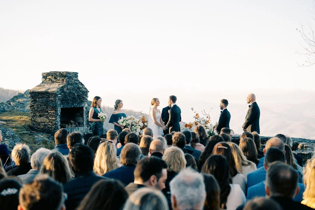 Wide shot of a mountain-top wedding ceremony with guests seated and the couple at the altar near a stone structure.