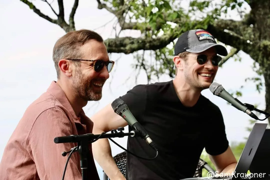 Two men, appearing to be in their 30s or 40s, are laughing while speaking into microphones.