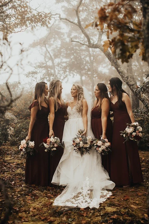 Bride standing with bridesmaids in burgundy dresses holding bouquets in a fall woodland setting.