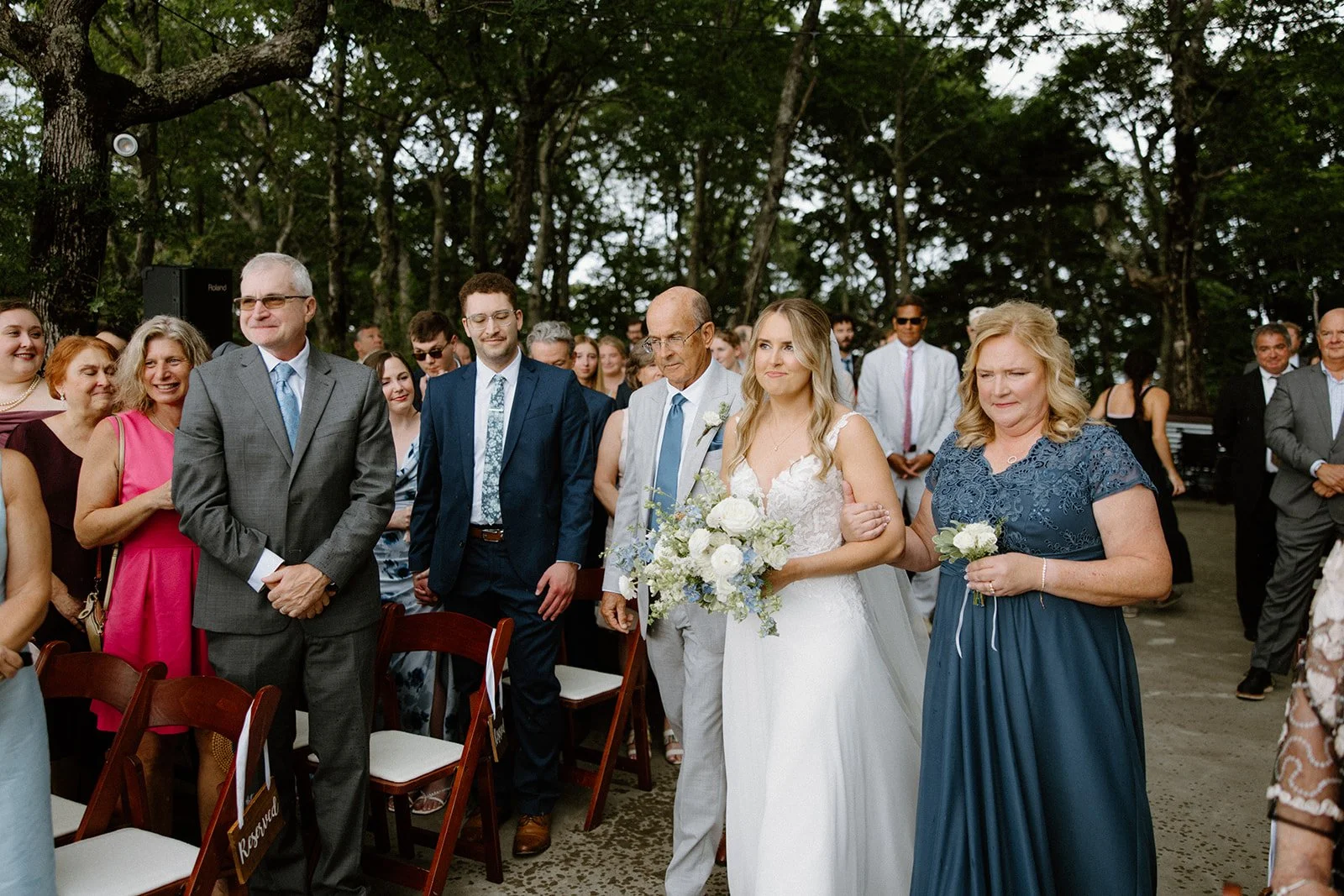 Bride walking down the aisle with her parents during an outdoor wedding ceremony surrounded by guests.