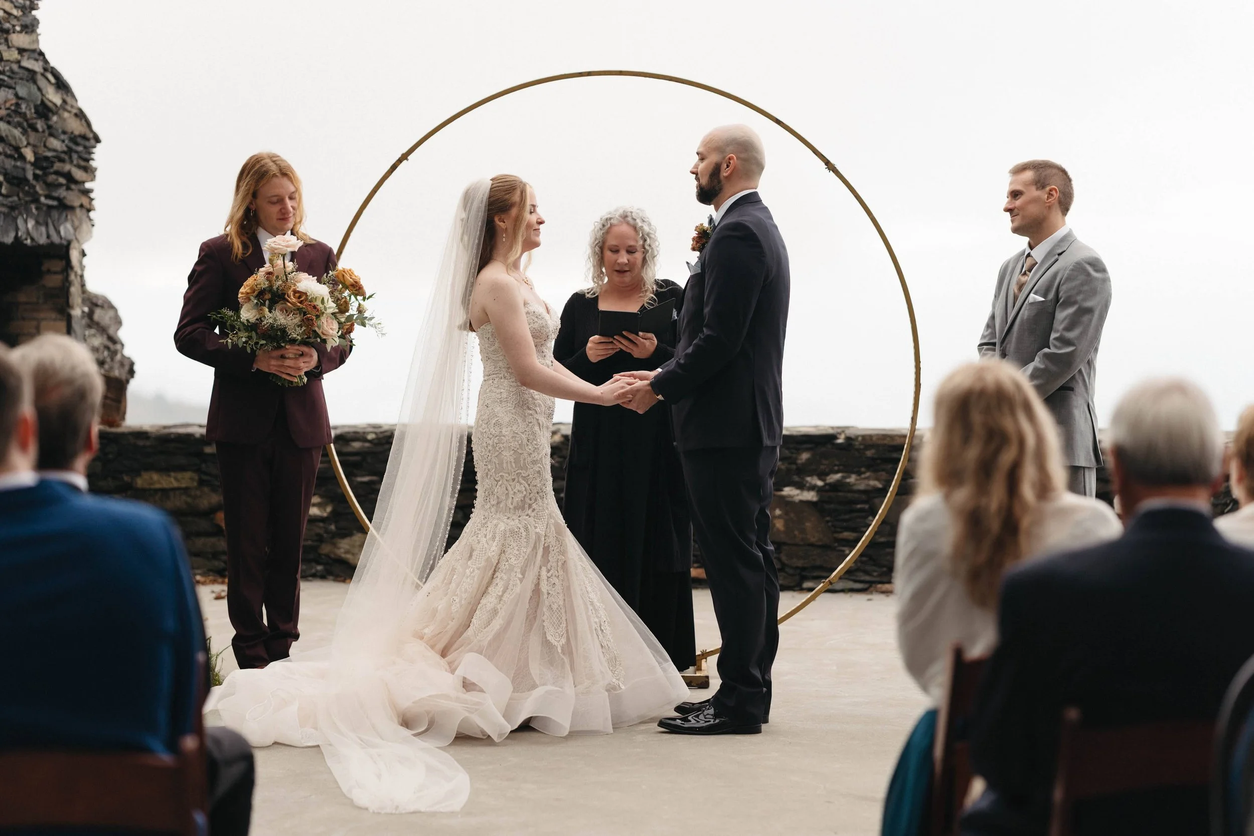 Bride and groom exchange vows in front of a large gold circular arch during an outdoor ceremony with guests seated nearby.