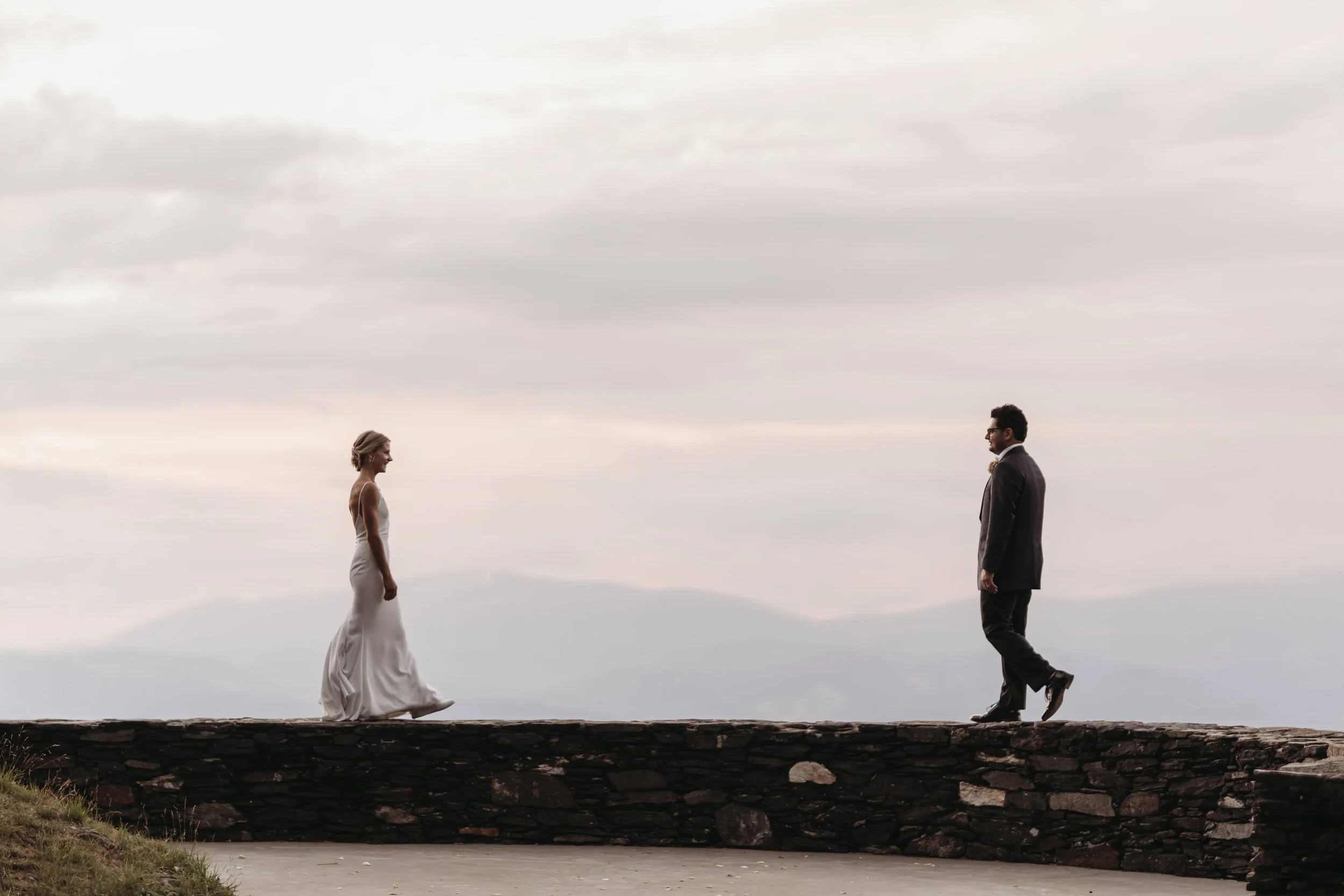 Bride and groom walking toward each other for a first look on a stone wall with soft mountain views behind them.