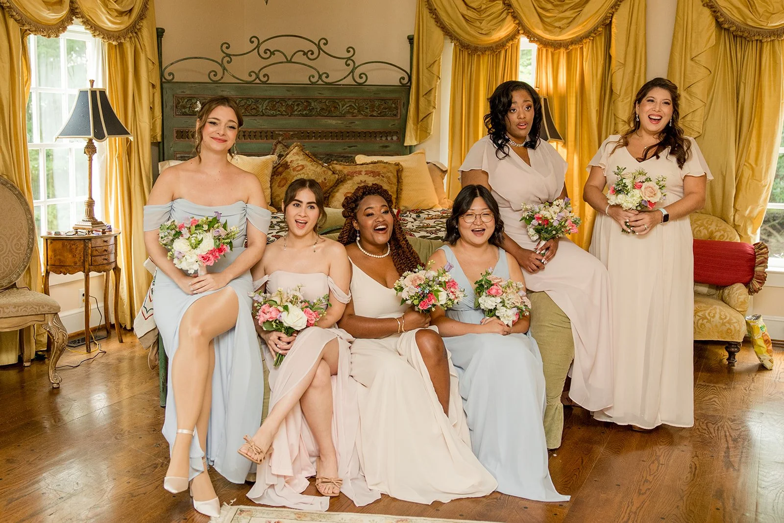 Bride posing with bridesmaids in pastel dresses while holding colorful floral bouquets in an elegant getting ready room.