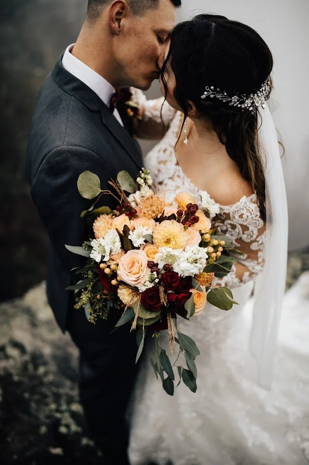 Closeup of the couple kissing with a fall floral bouquet and the bride’s lace sleeves visible.