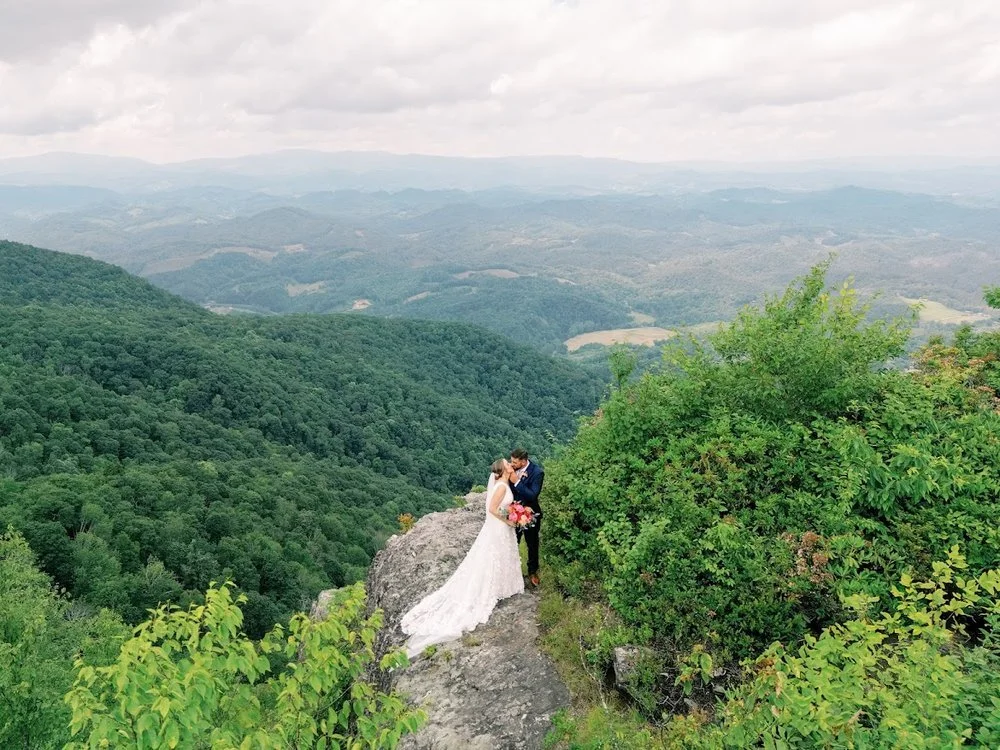 A newlywed couple embraces on a rocky mountain overlook with vast green mountains stretching into the distance.