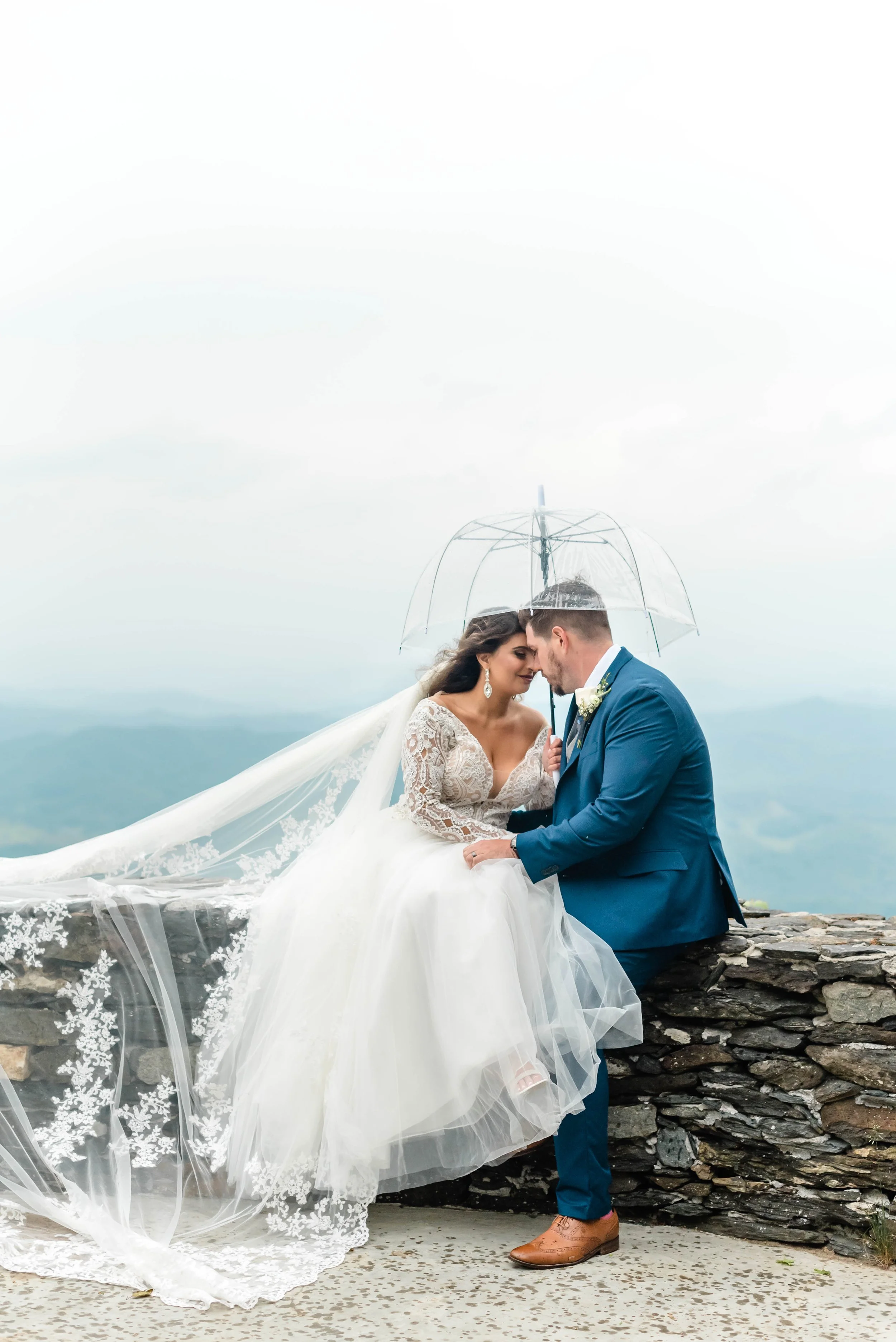 Bride and groom sitting together under a clear umbrella at a misty mountain overlook.