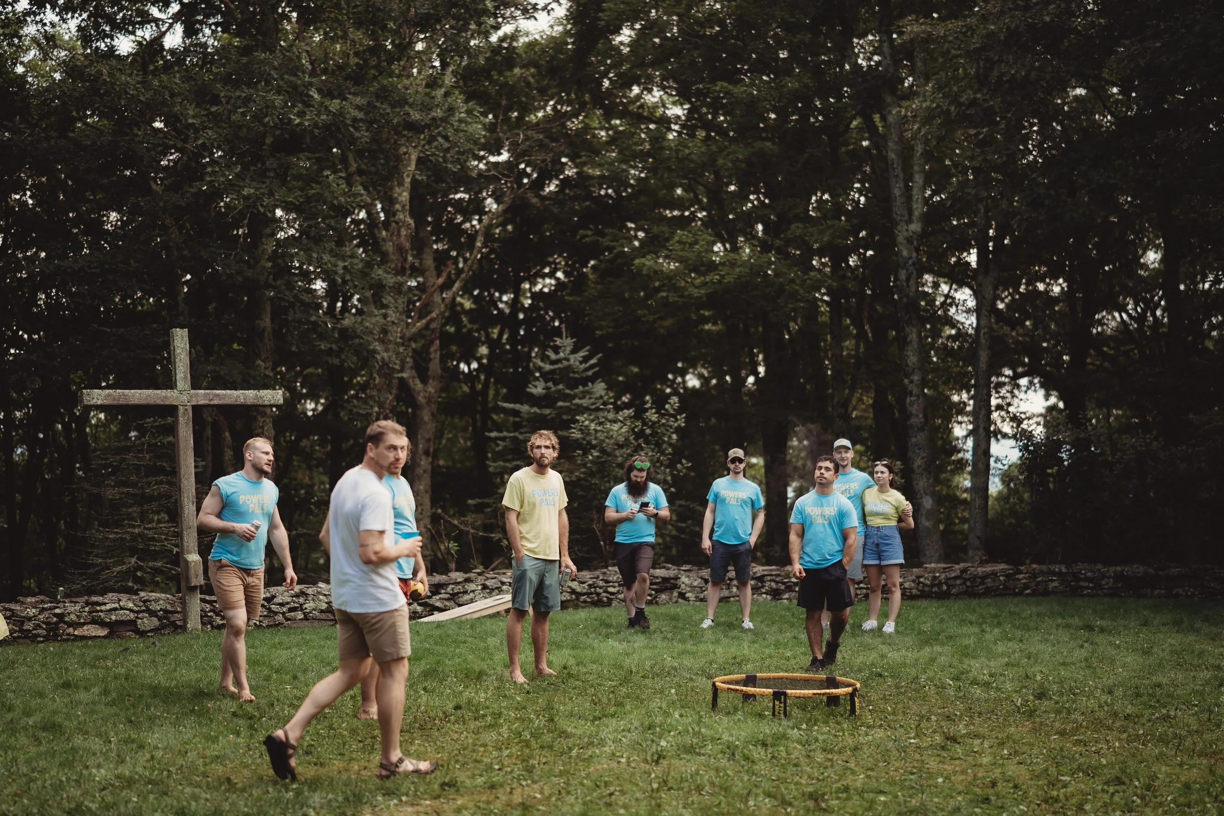 Guests playing a casual lawn game during a wedding weekend event near a wooden cross, stone wall, and wooded backdrop.