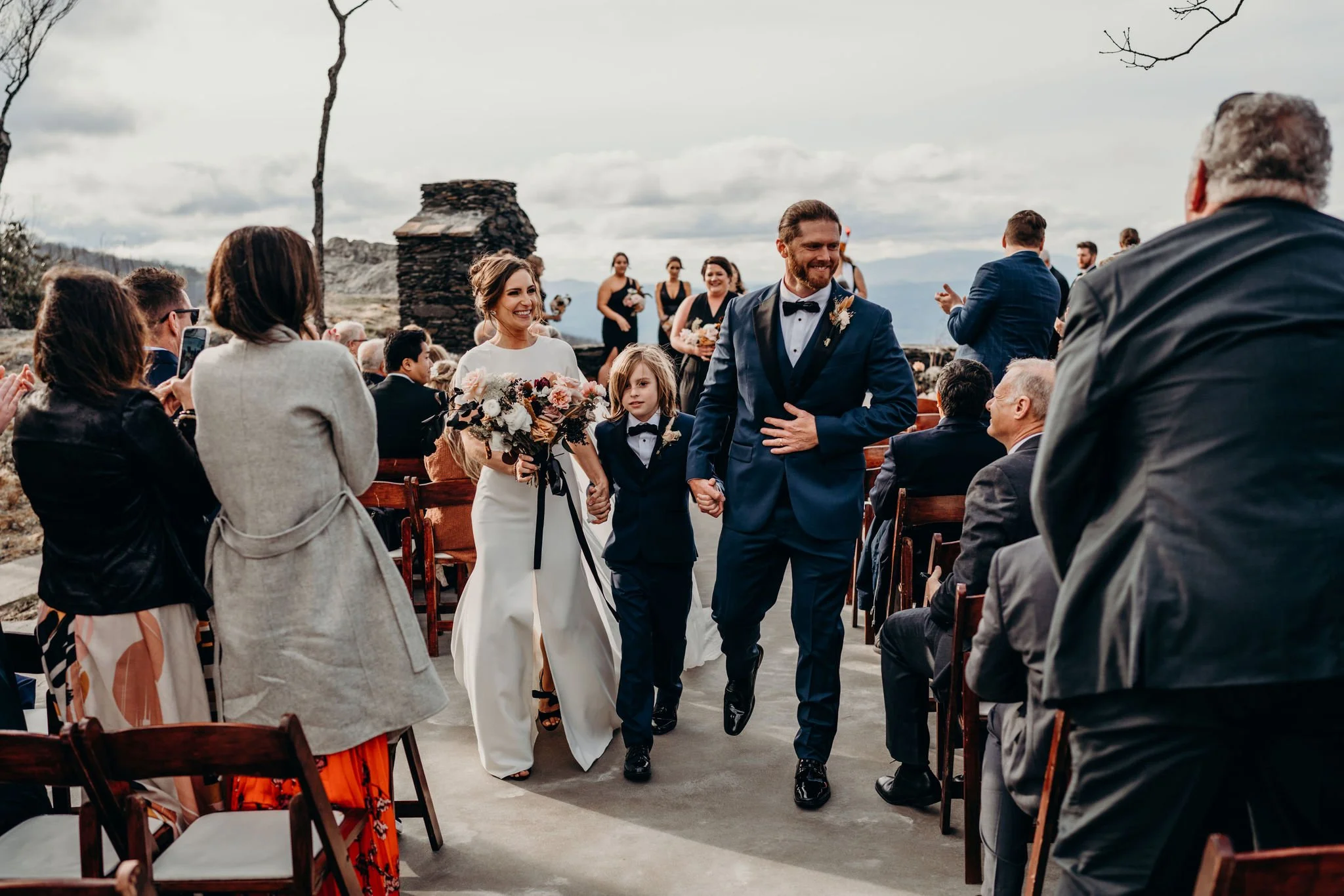 Couple and child walking up the aisle after an outdoor mountain ceremony as guests watch.