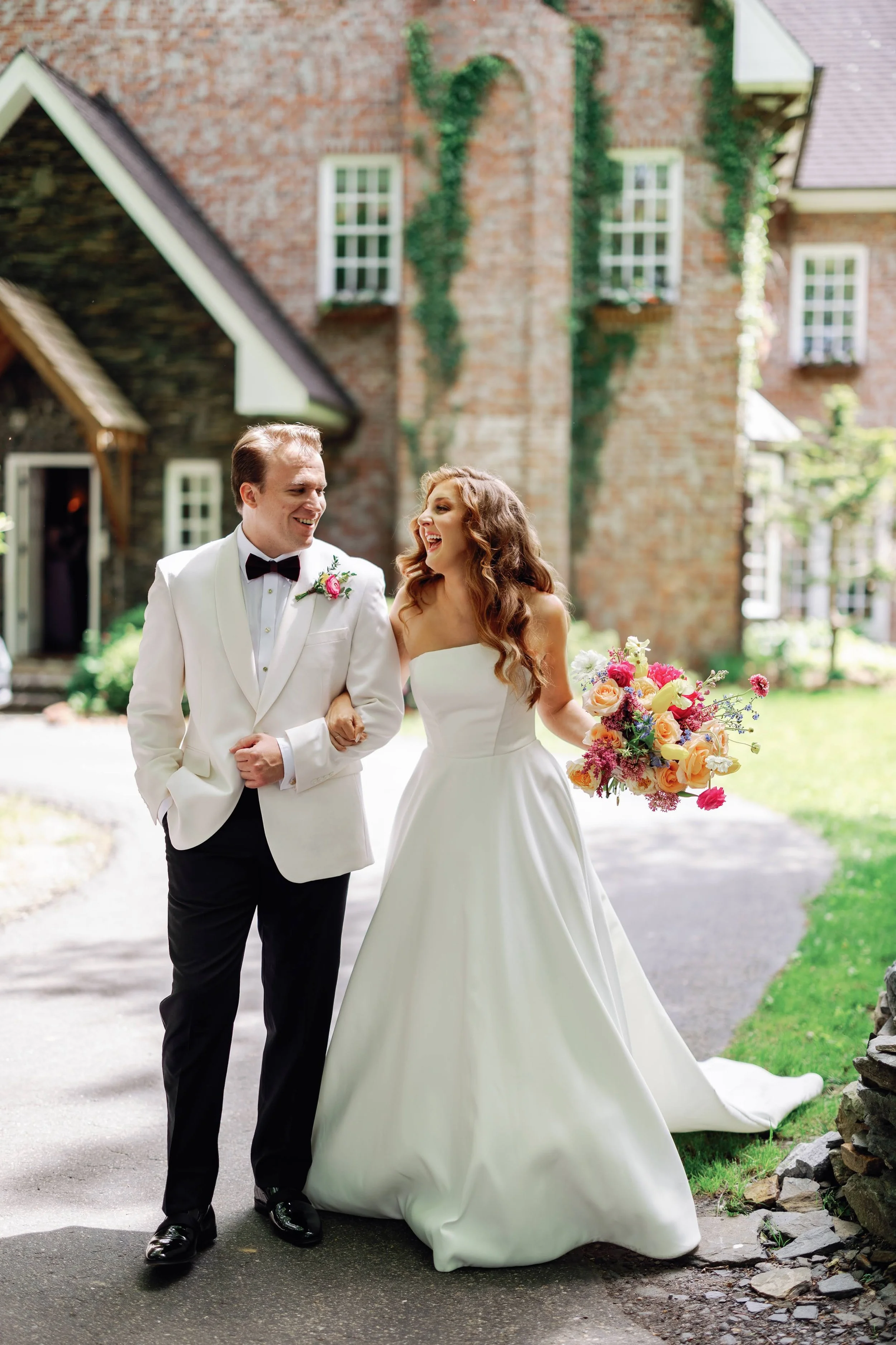 Bride and groom walk arm in arm outside a brick estate building, smiling and holding a bright colorful bouquet.