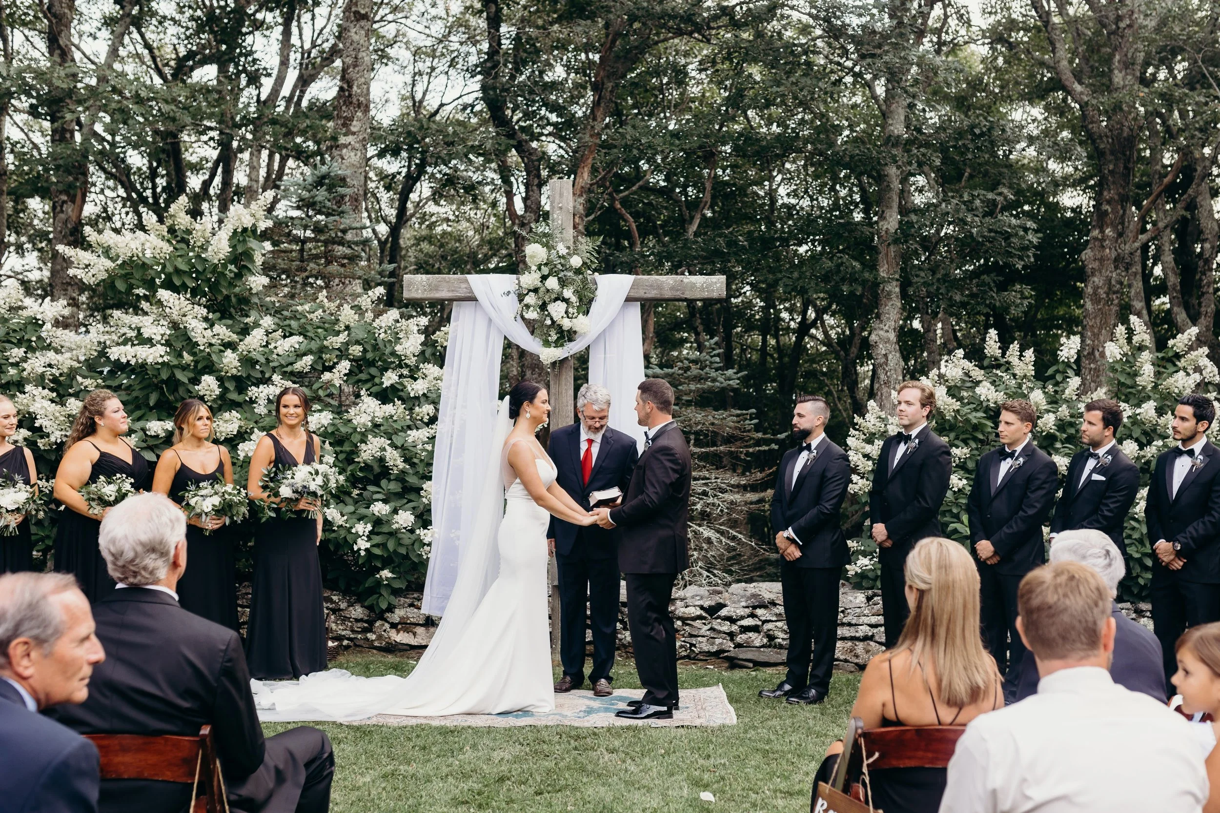 Garden wedding ceremony with a wooden arbor draped in white fabric as the couple holds hands and the wedding party stands nearby.
