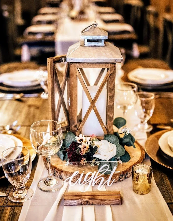 Rustic wedding tablescape with wooden lantern, eucalyptus, candlelight, and “one” table number on a long banquet table.