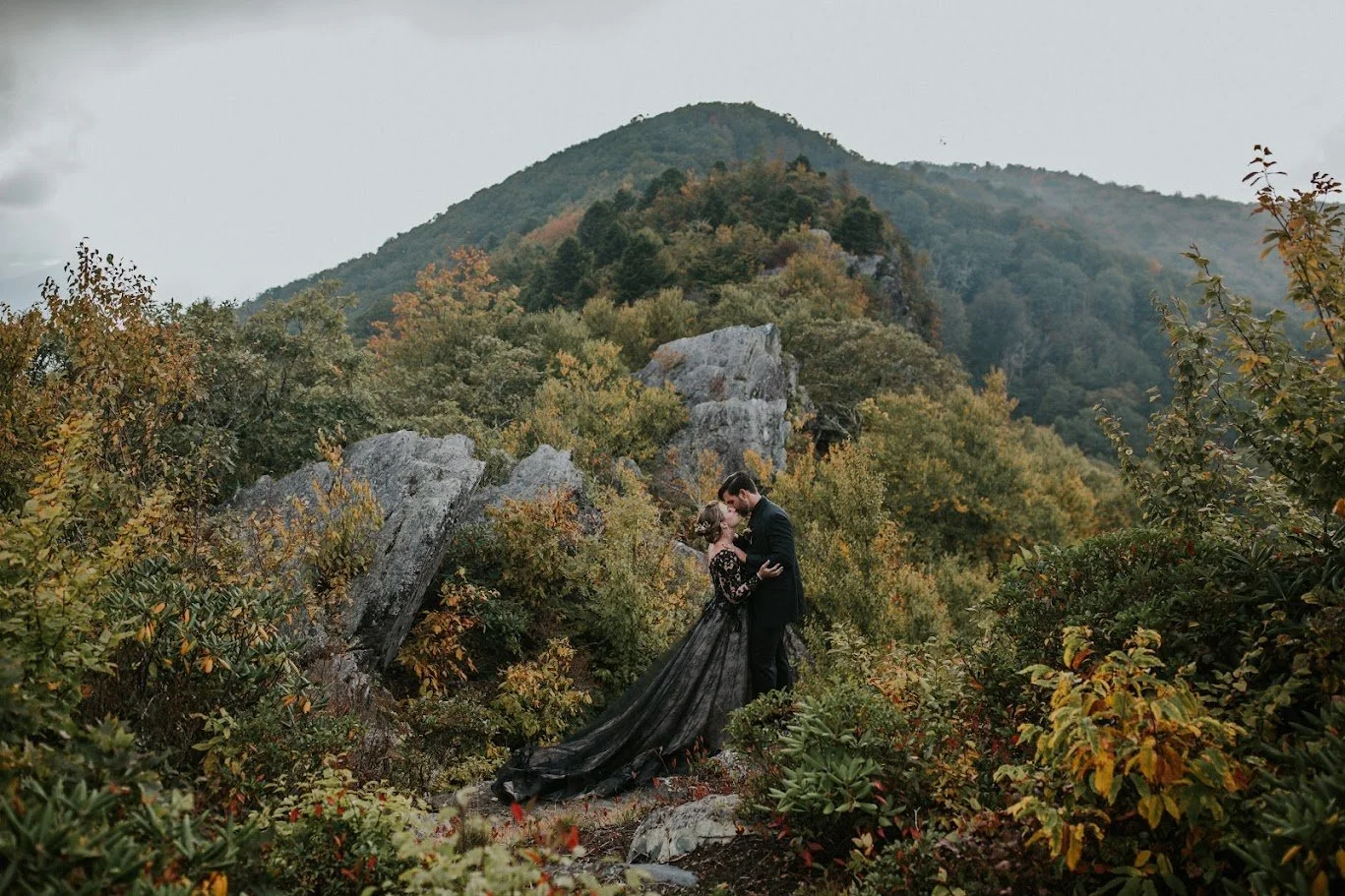 Couple kissing on a mountain trail surrounded by fall foliage, bride wearing a long black lace wedding dress.