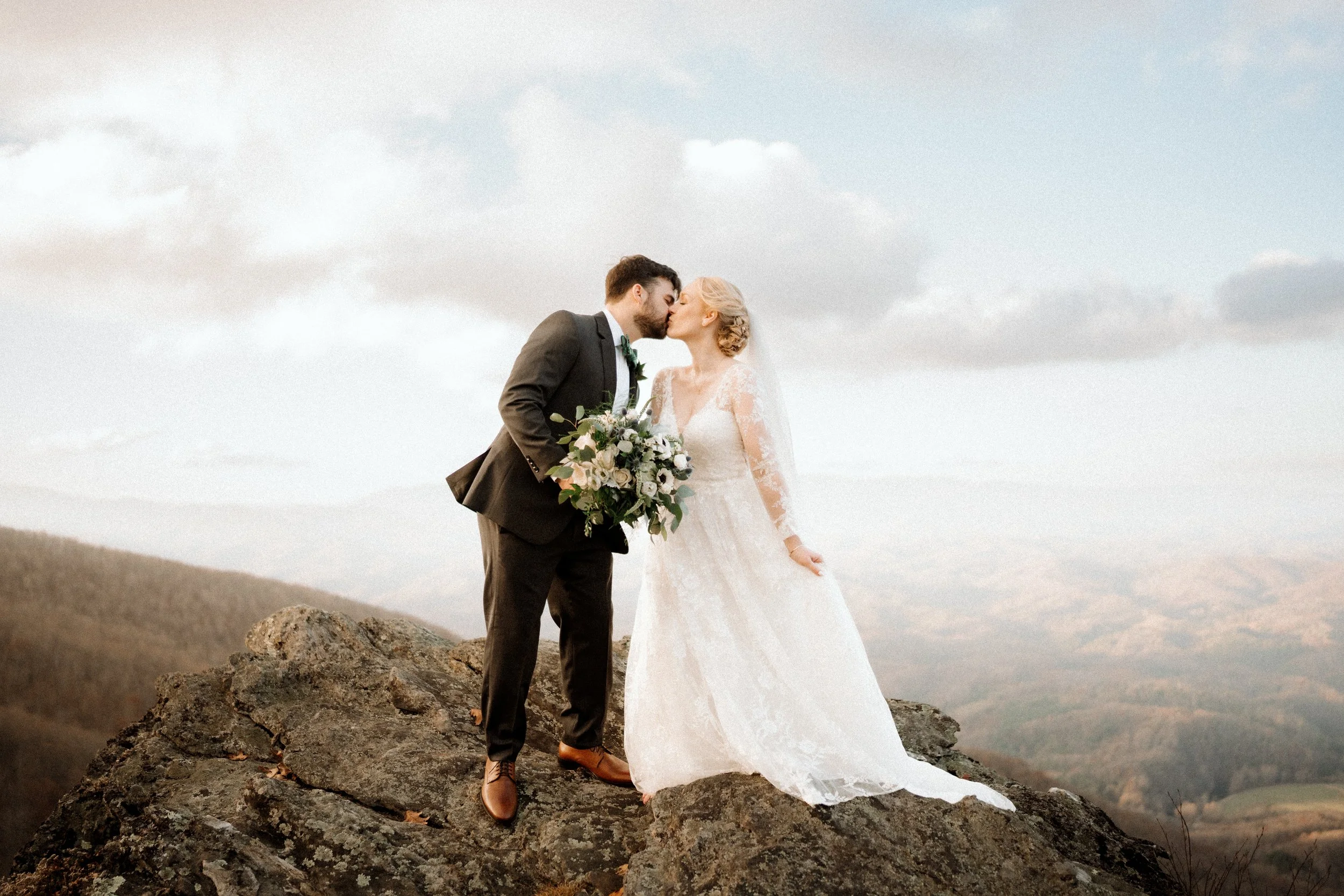 Bride and groom kissing on a rocky mountaintop with a dramatic sky and sweeping mountain views