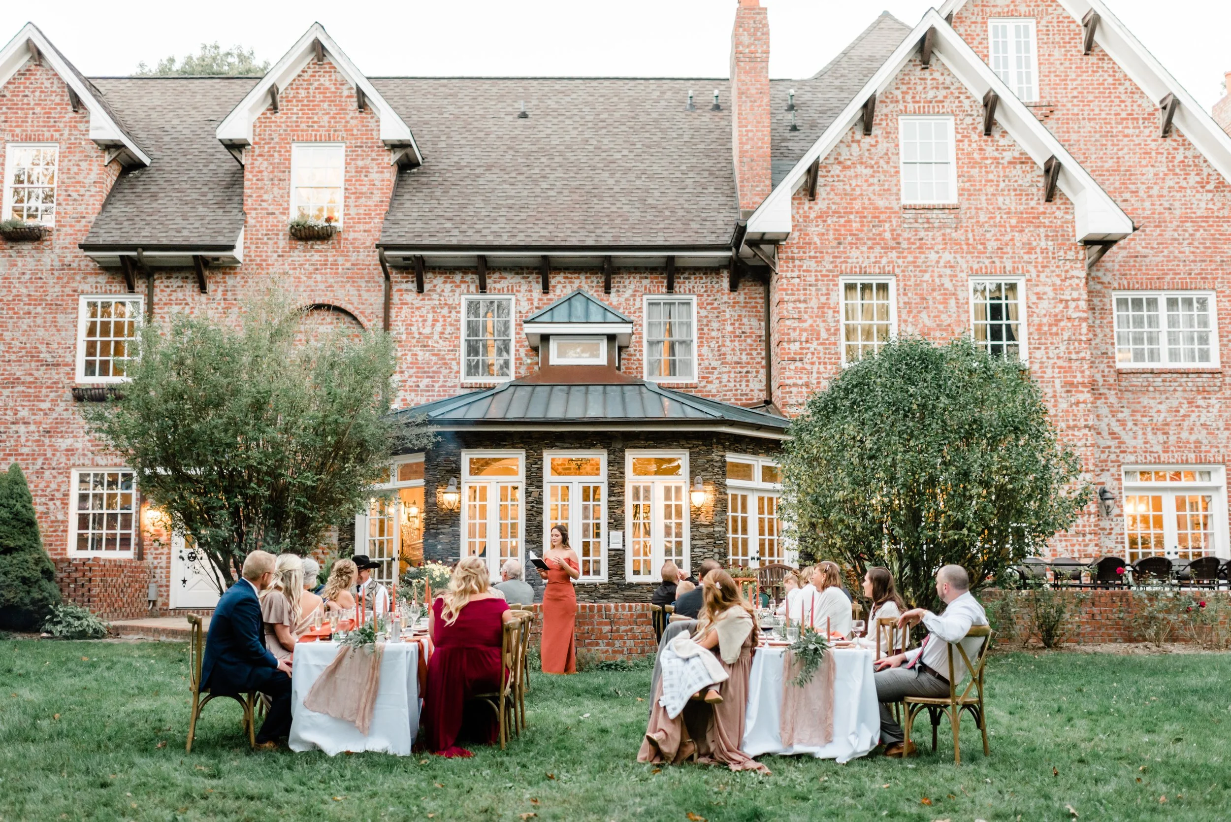 Garden wedding reception outside a historic brick estate with round tables, greenery, and guests dining on the lawn.