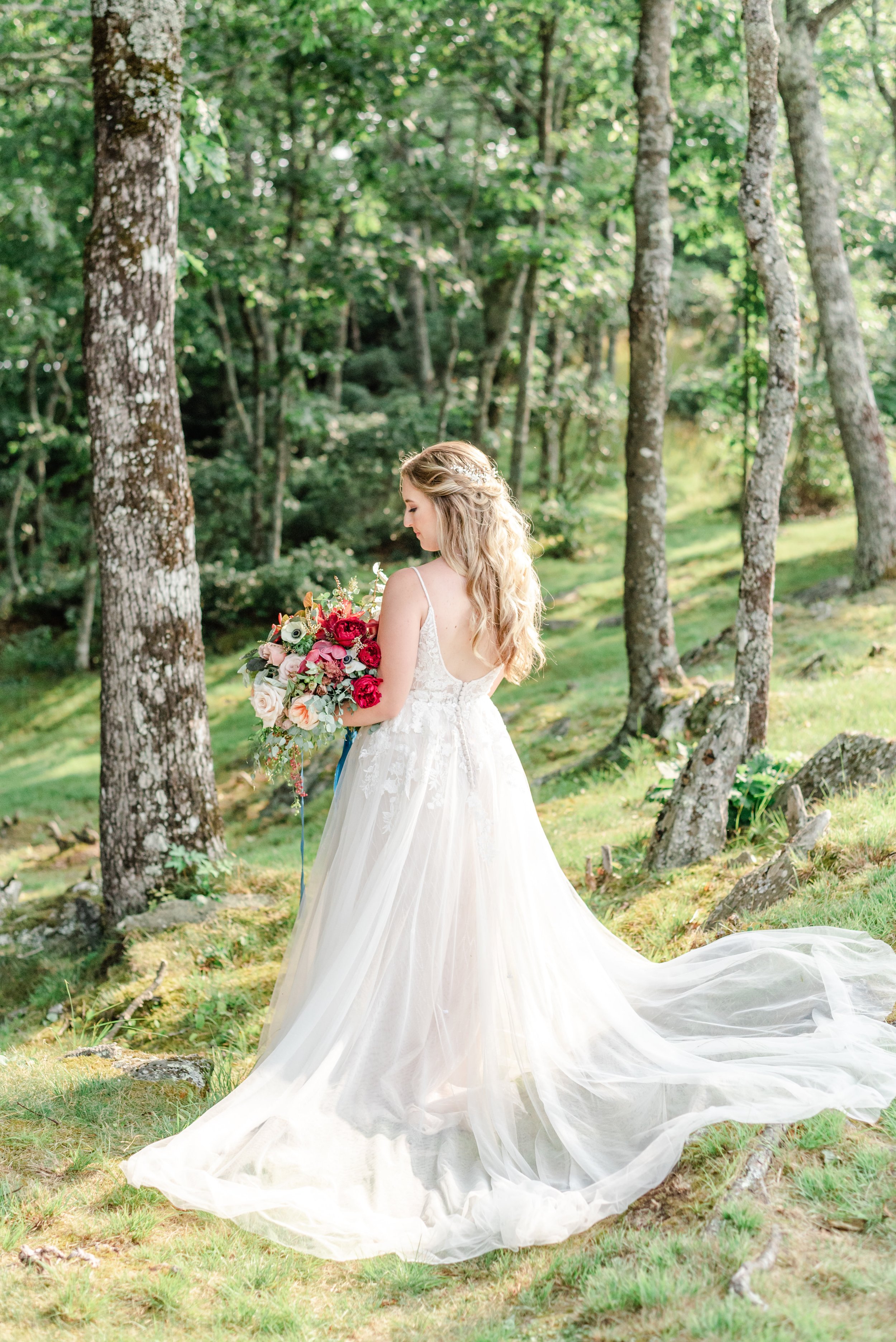 Back view of the bride holding a bouquet with a long train flowing on a green forest hillside.