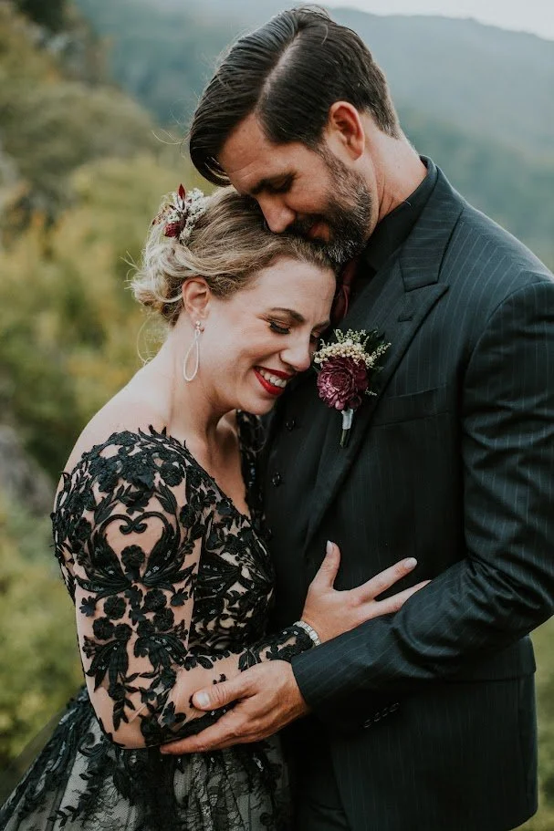 Closeup of the couple embracing, bride in black lace gown and groom in a dark suit with boutonniere.