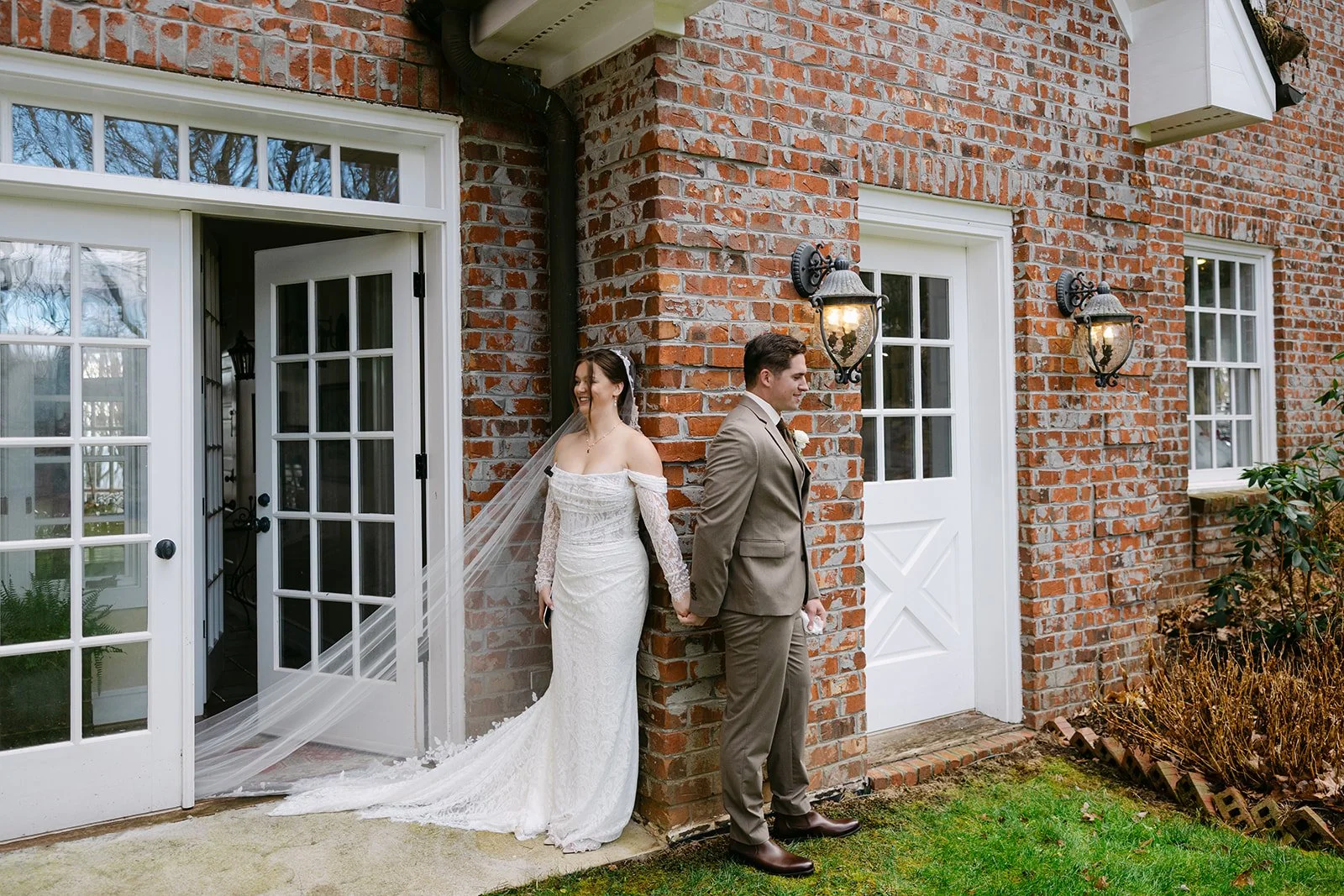 Bride and groom holding hands outside a brick wedding venue with white doors and vintage wall lanterns.