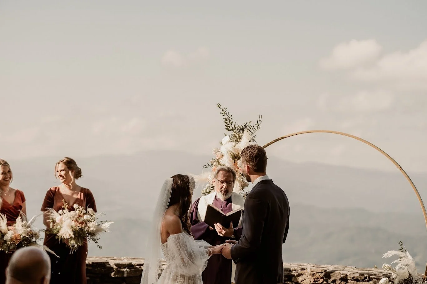 Bride and groom exchanging vows under a circular floral arch on a mountaintop with mountains in the background