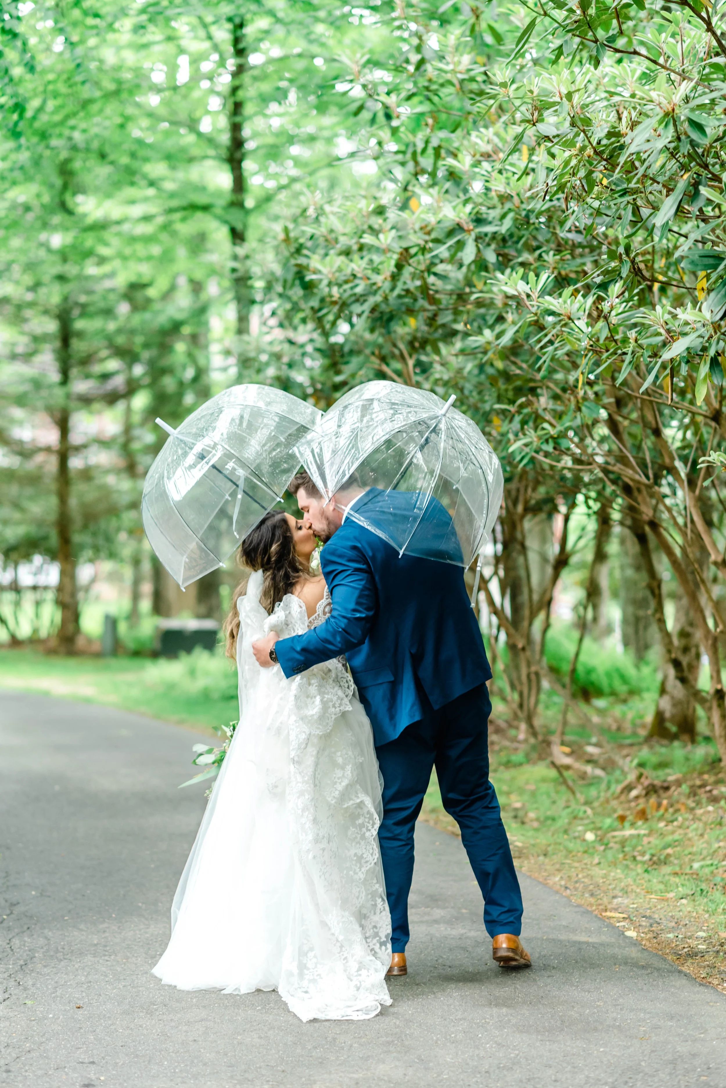 Bride and groom kissing under clear umbrellas on a wooded path after their wedding ceremony.