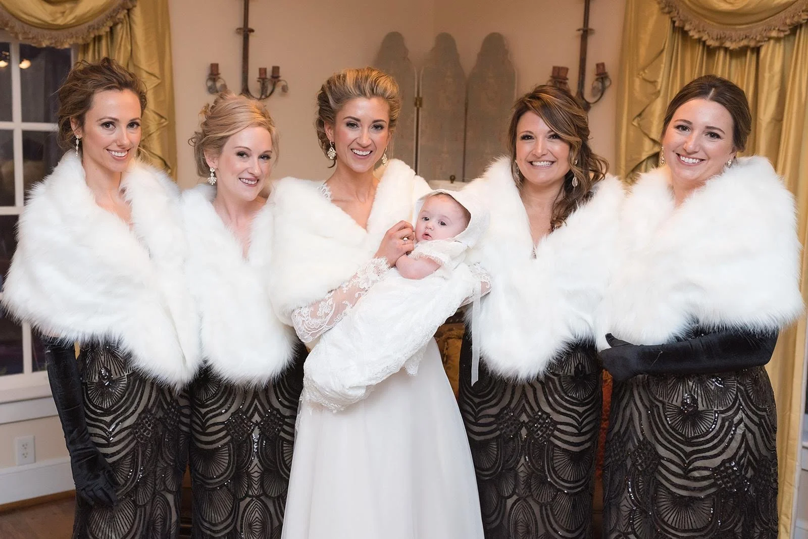 Bride and bridesmaids in fur wraps posing indoors while holding a baby.