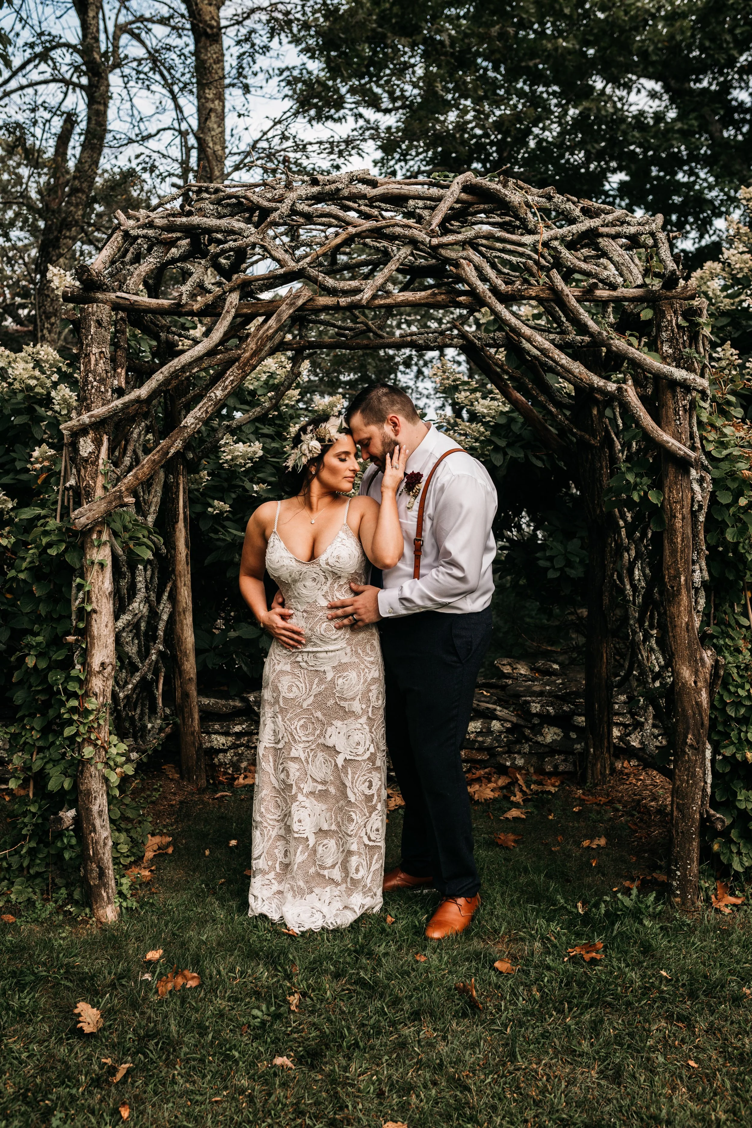 Bride and groom embracing under a rustic branch arbor in a wooded outdoor setting.
