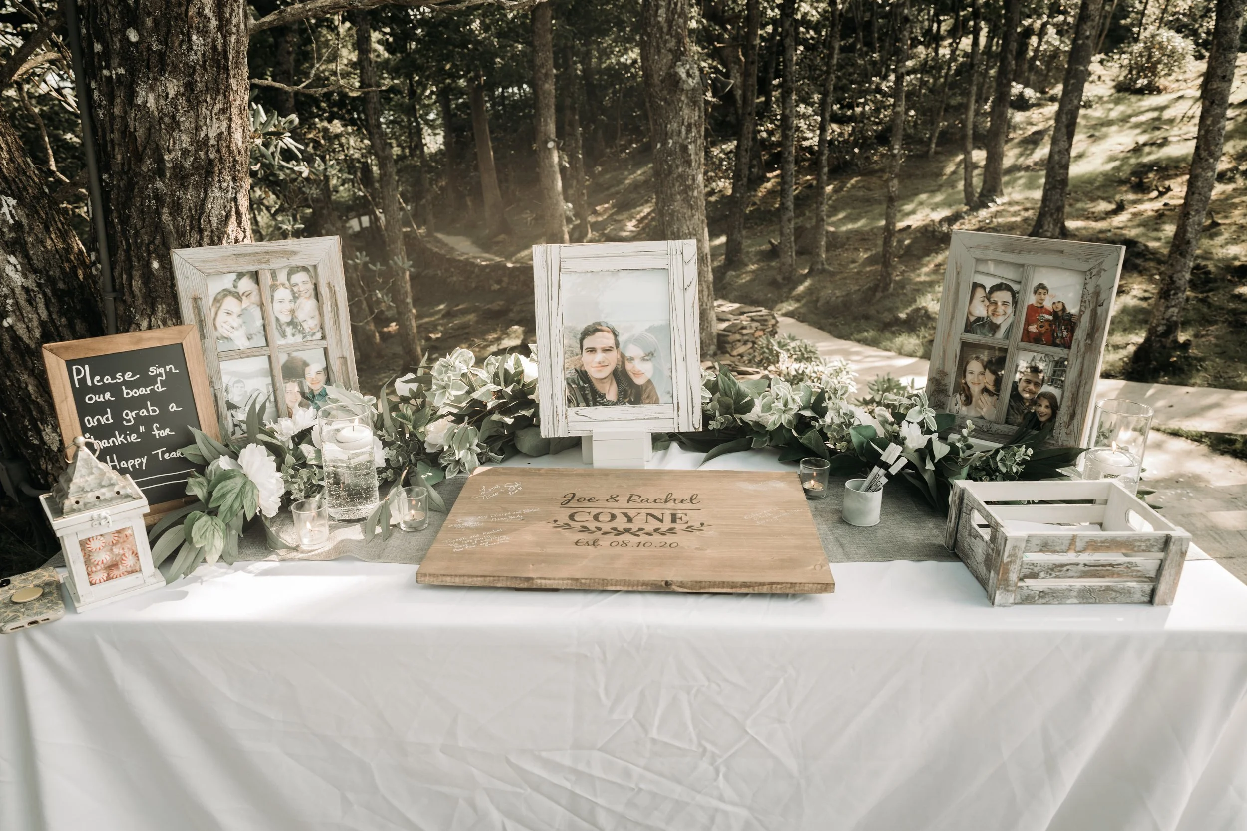 Wedding memory table with framed family photos, candles, greenery, and personalized wooden sign outdoors.