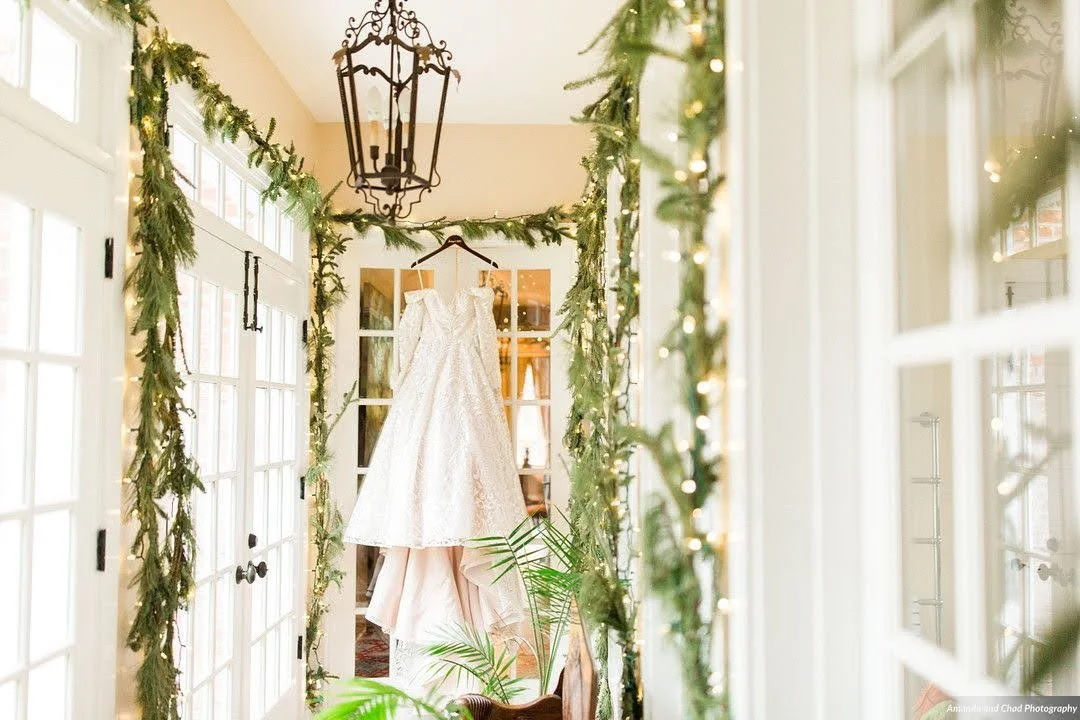 Wedding dress hanging in a bright hallway decorated with greenery garlands and lantern lighting.