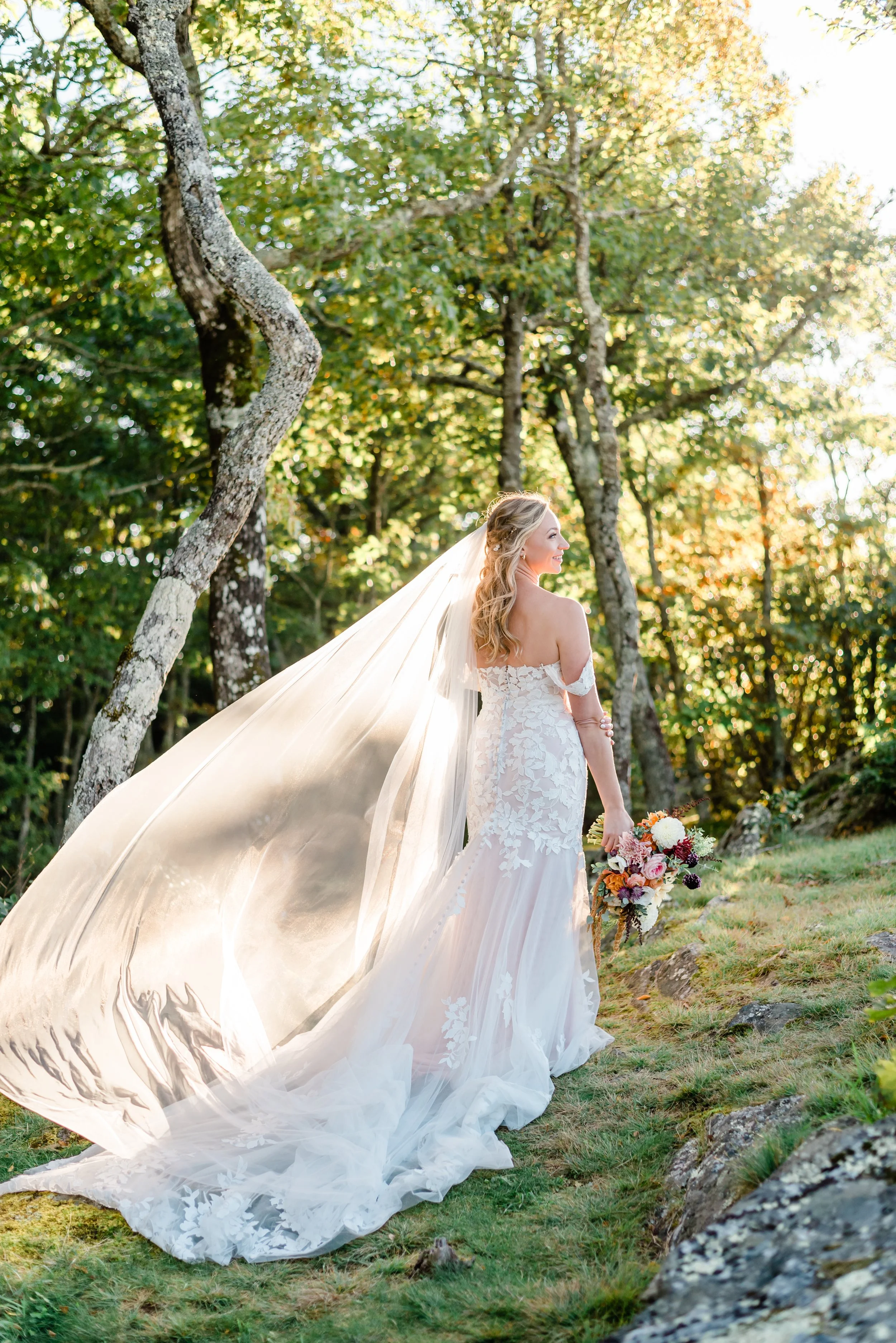 Bride standing in a forest clearing with her long veil blowing in the sunlight while holding a bouquet