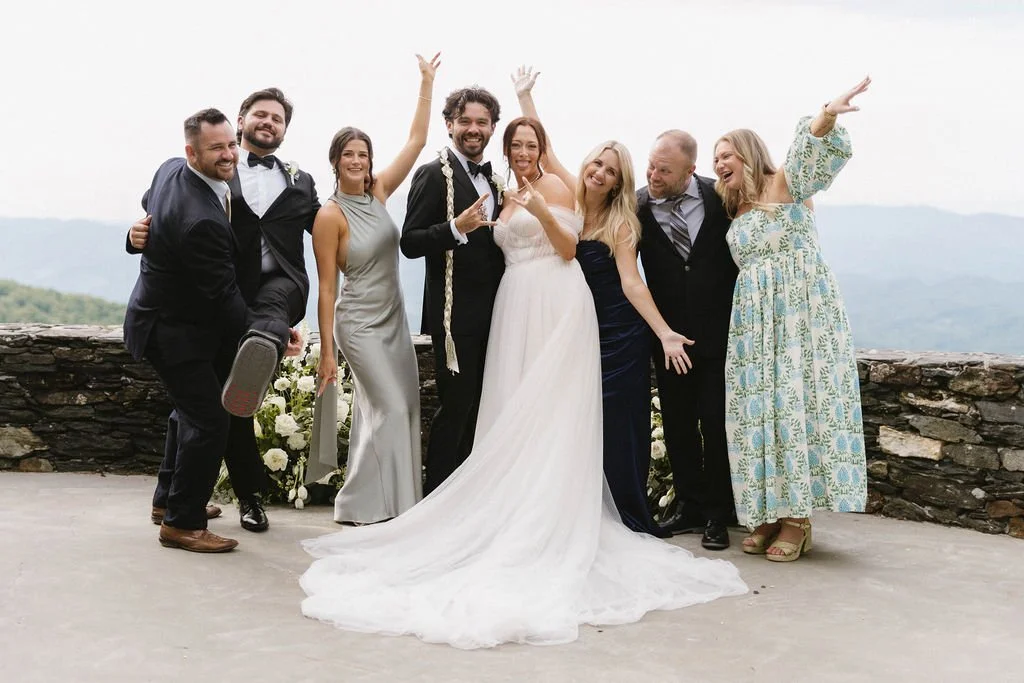 Bride and groom posing with a joyful wedding party at a scenic mountain overlook with guests cheering beside them.