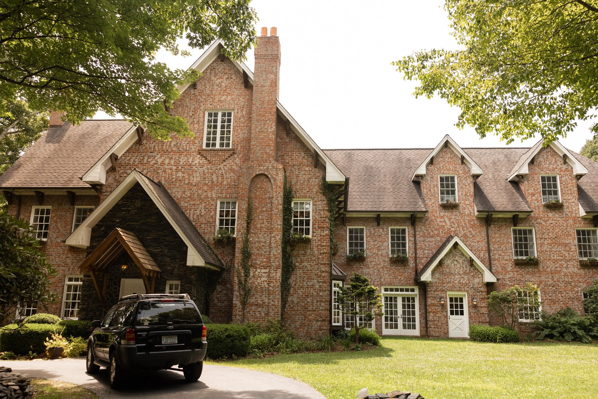 Exterior view of a large brick estate building with a driveway and parked car surrounded by trees.