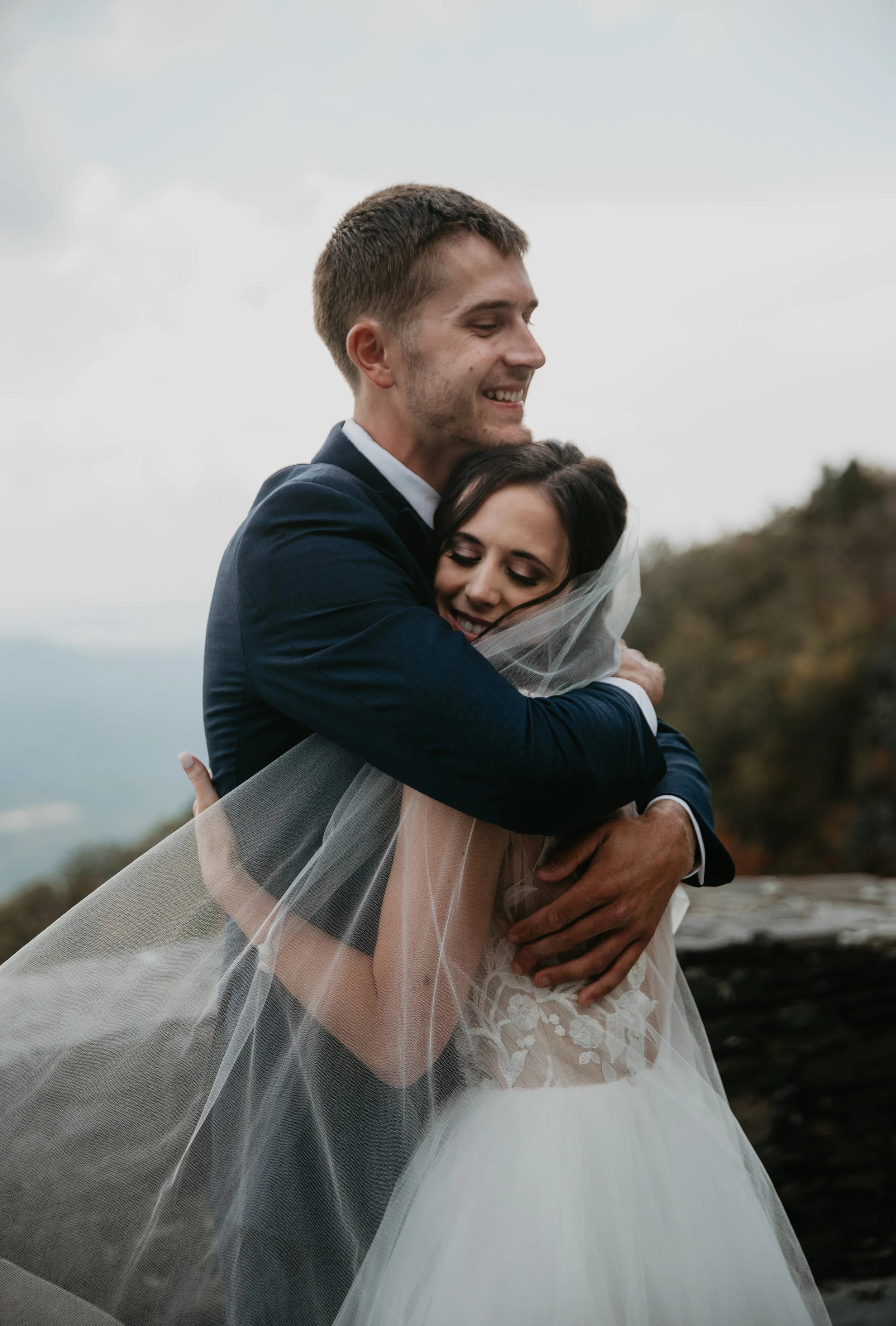 Groom hugs bride in a veil on a mountain overlook during an outdoor wedding portrait.