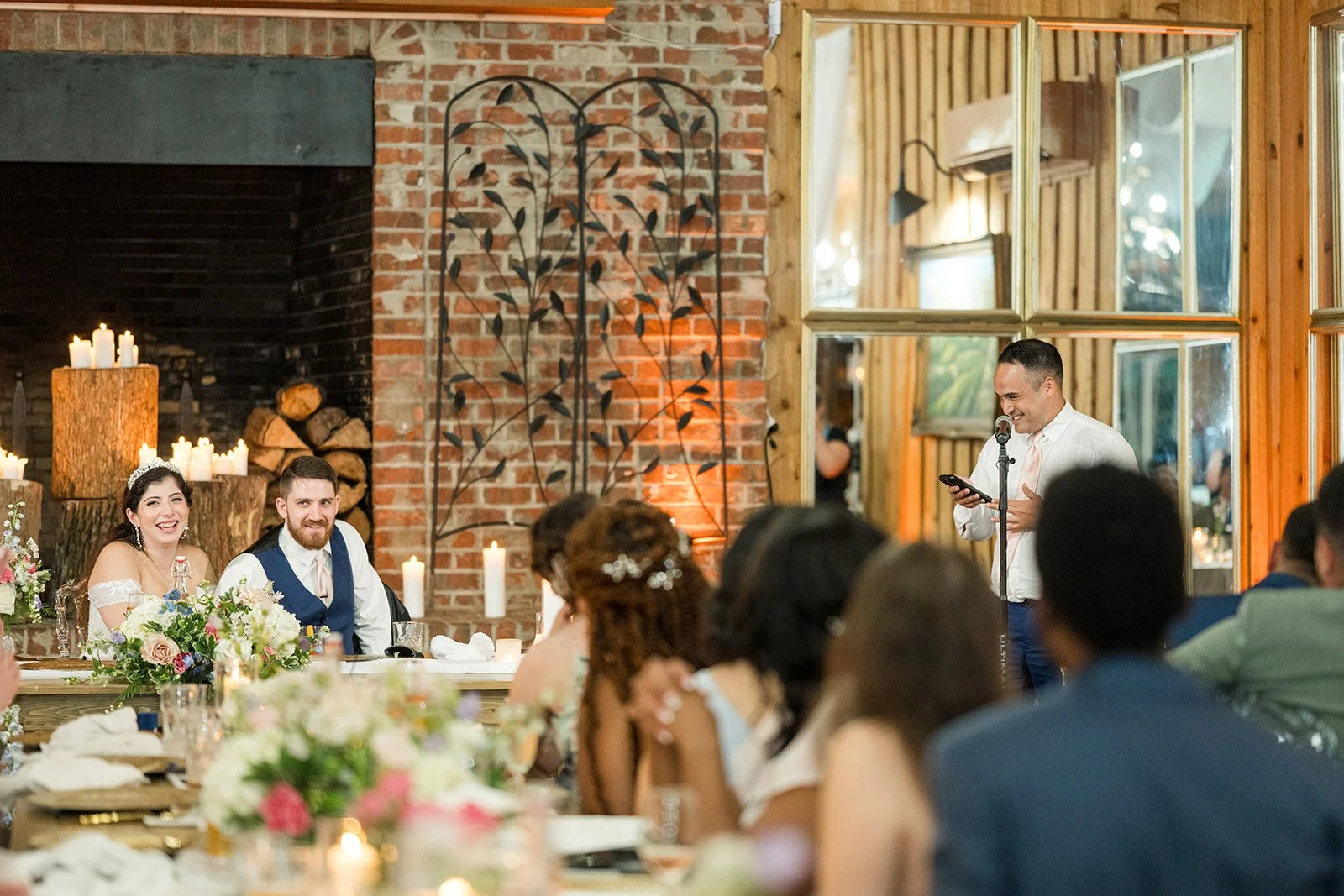 Groomsman giving a wedding reception speech while the bride and groom smile at their sweetheart table.