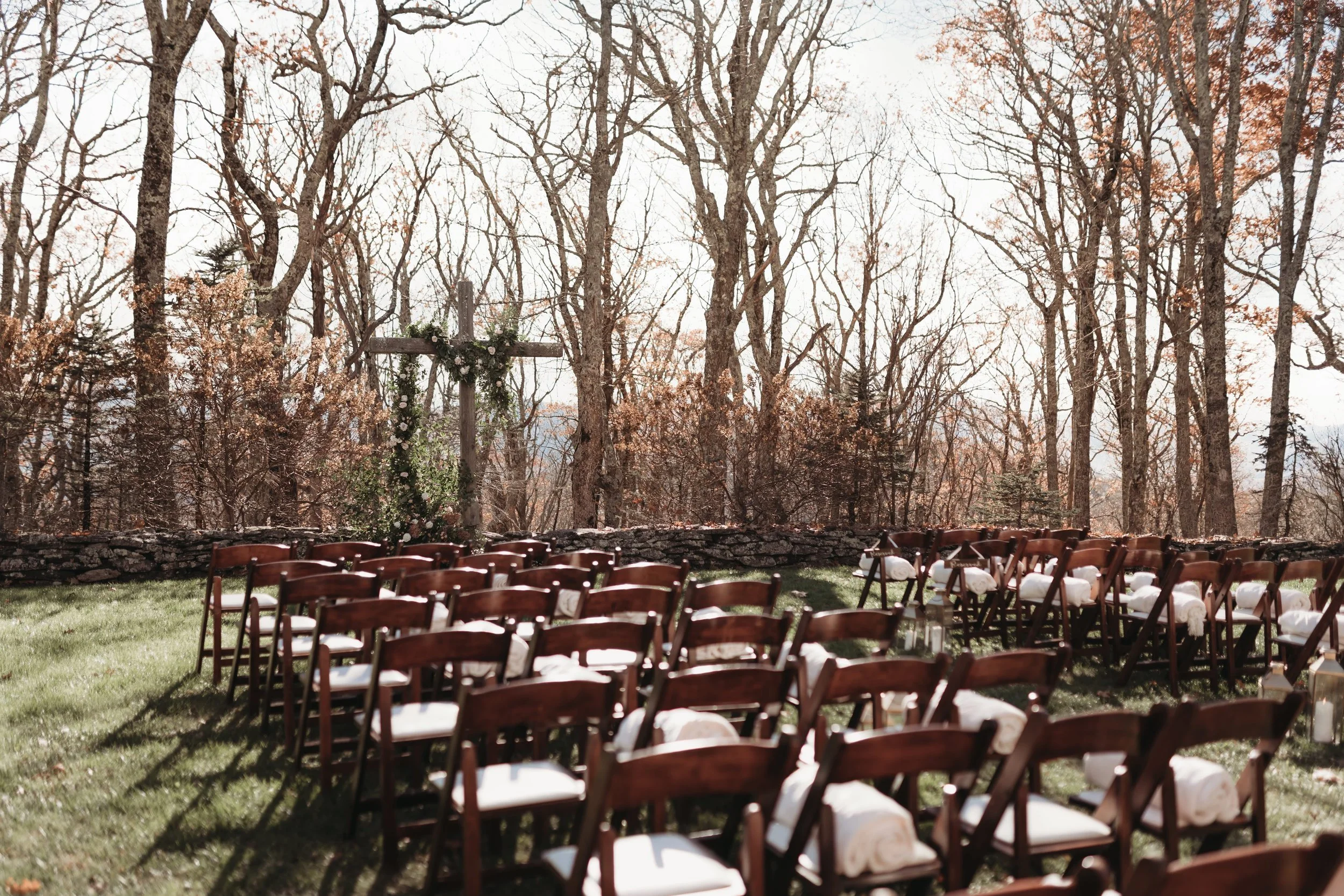 Outdoor woodland wedding ceremony setup with wooden chairs and a cross decorated with greenery
