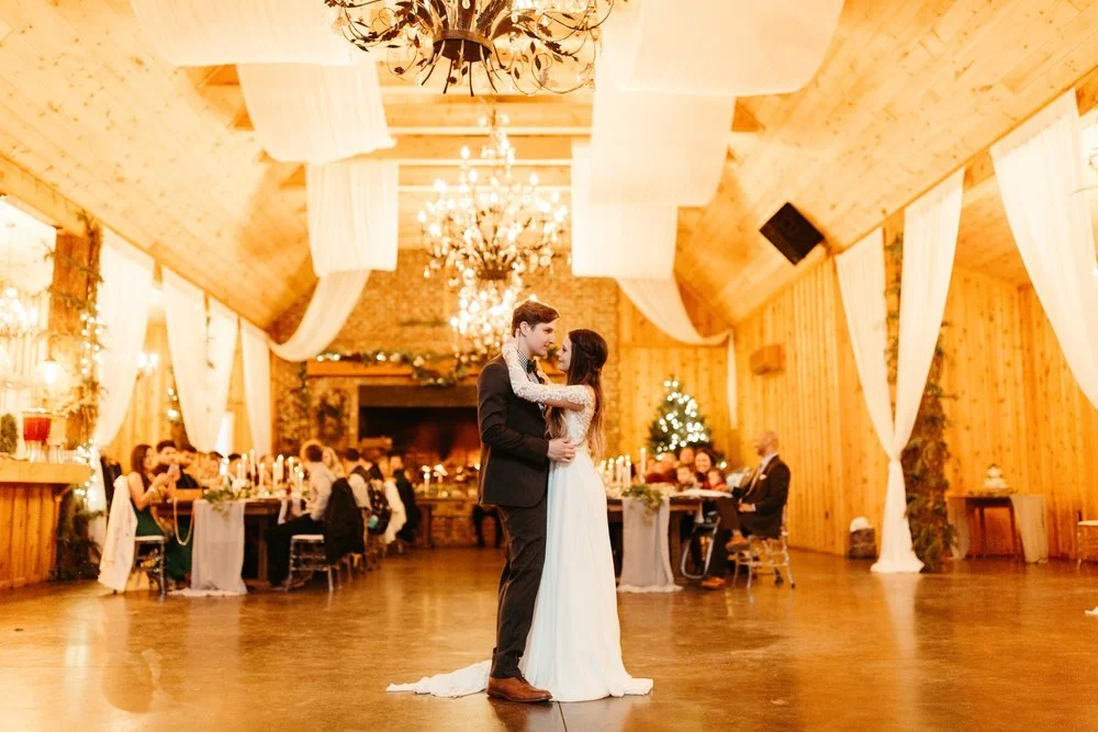 A newlywed couple embraces on a dance floor in a rustic venue with high ceilings and chandeliers, guests seated at tables.