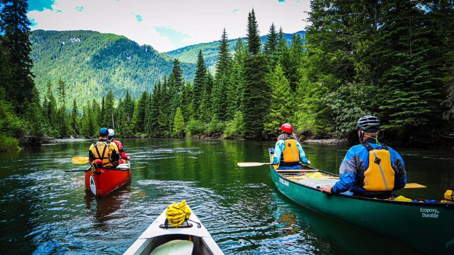 Canoes on a calm mountain river surrounded by evergreen forest and hills.