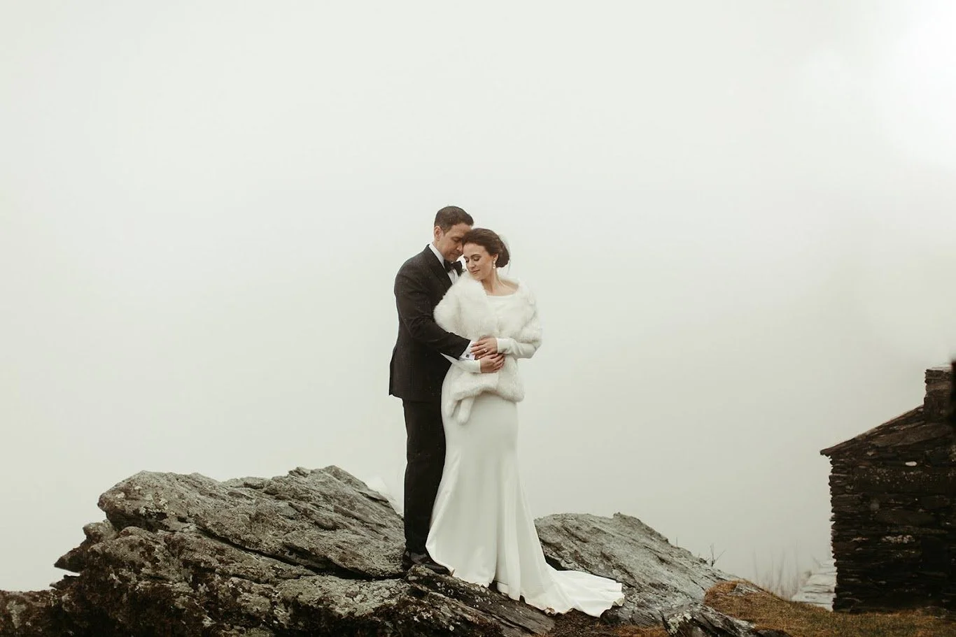 Bride and groom embracing on rocks in a foggy mountain landscape, wearing winter wedding attire.