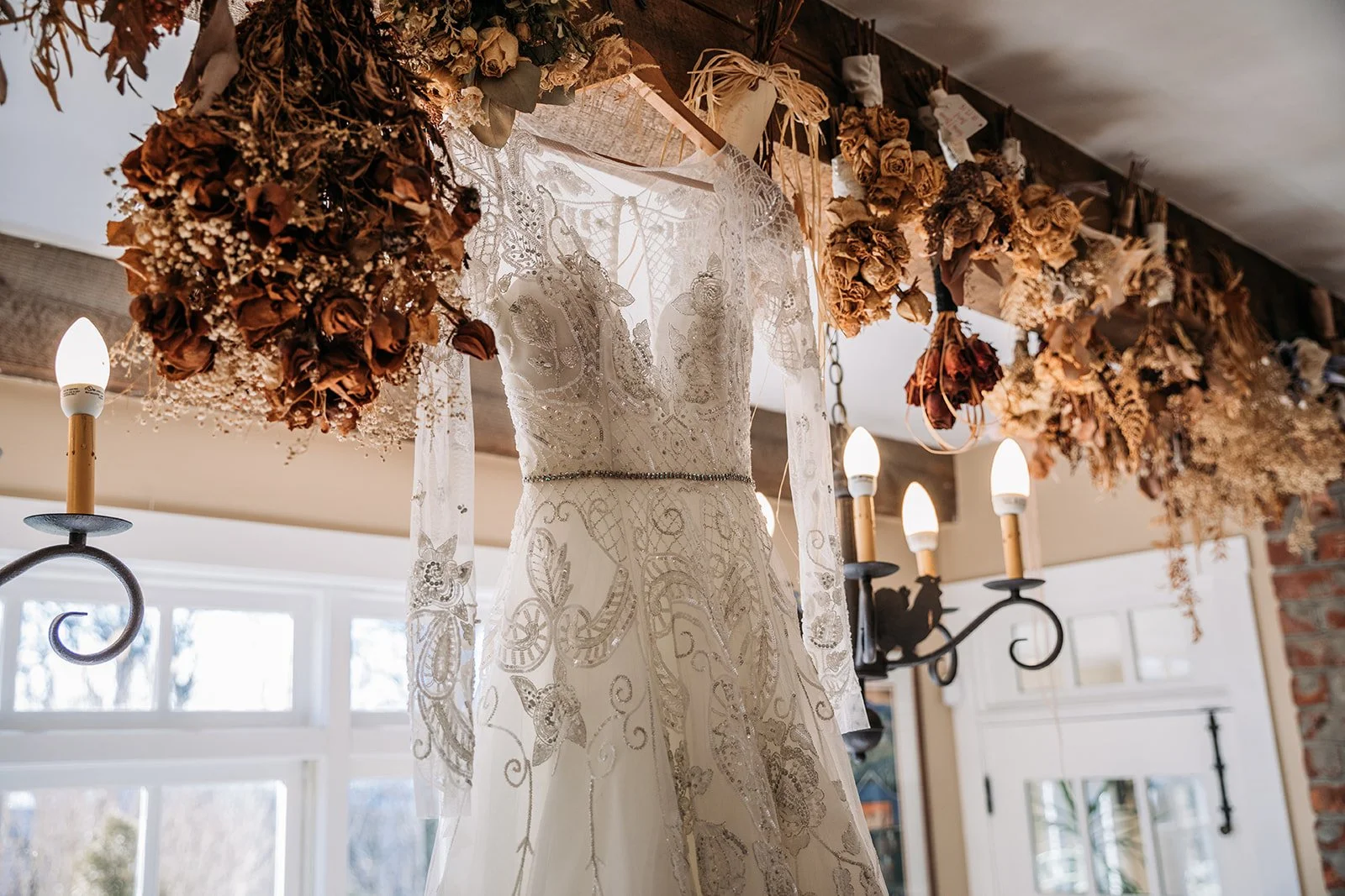 Beaded lace wedding dress hanging indoors with rustic dried floral decor around the hanger.