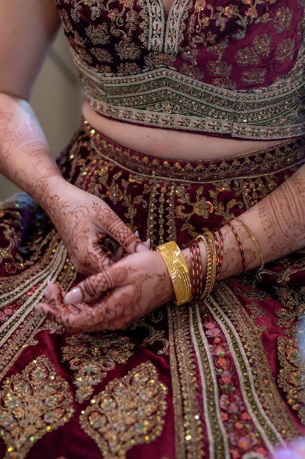 Close-up of hands with henna and bangles resting on an embroidered maroon outfit with gold detailing.